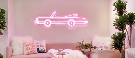 A pink-themed living room with a neon car sign, a sofa, a coffee table, and various plants.