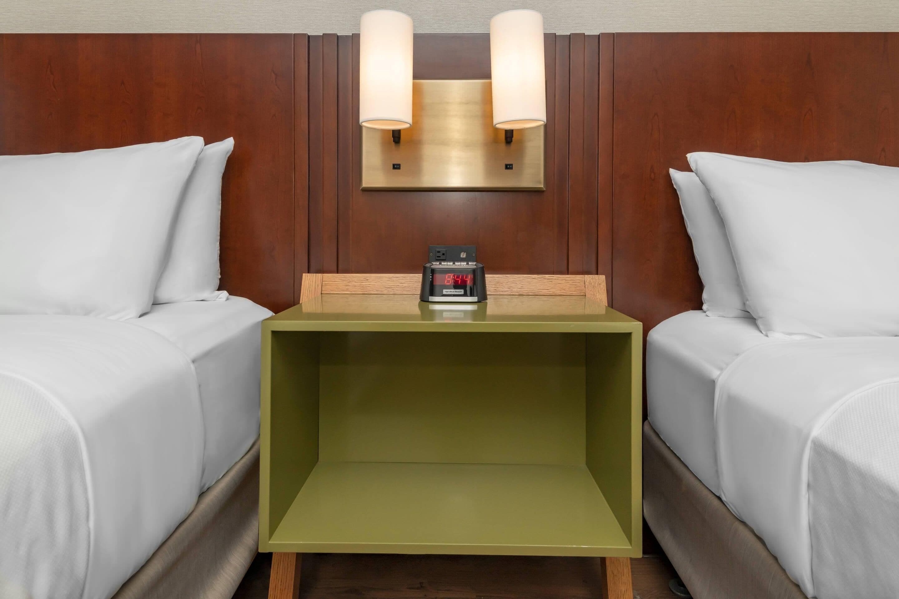 Habitación de hotel con dos camas, una mesita de noche con reloj digital y lámparas de pared.