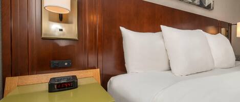 Une chambre d’hôtel avec une tête de lit en bois, un lit avec une literie blanche, une table de chevet avec une horloge et une lampe fixée au mur.