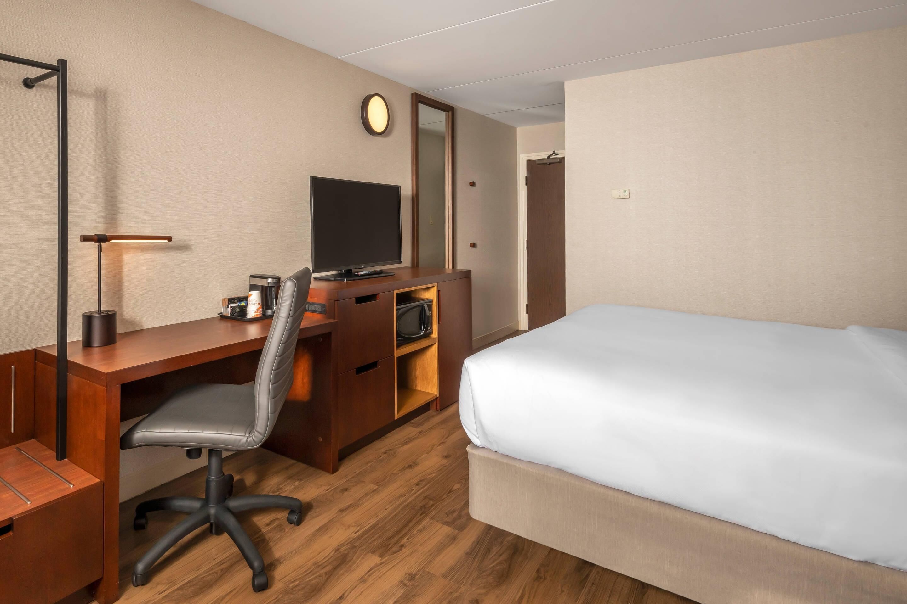 A hotel room with a wooden desk, a television, a chair, a bed, and a mirror.