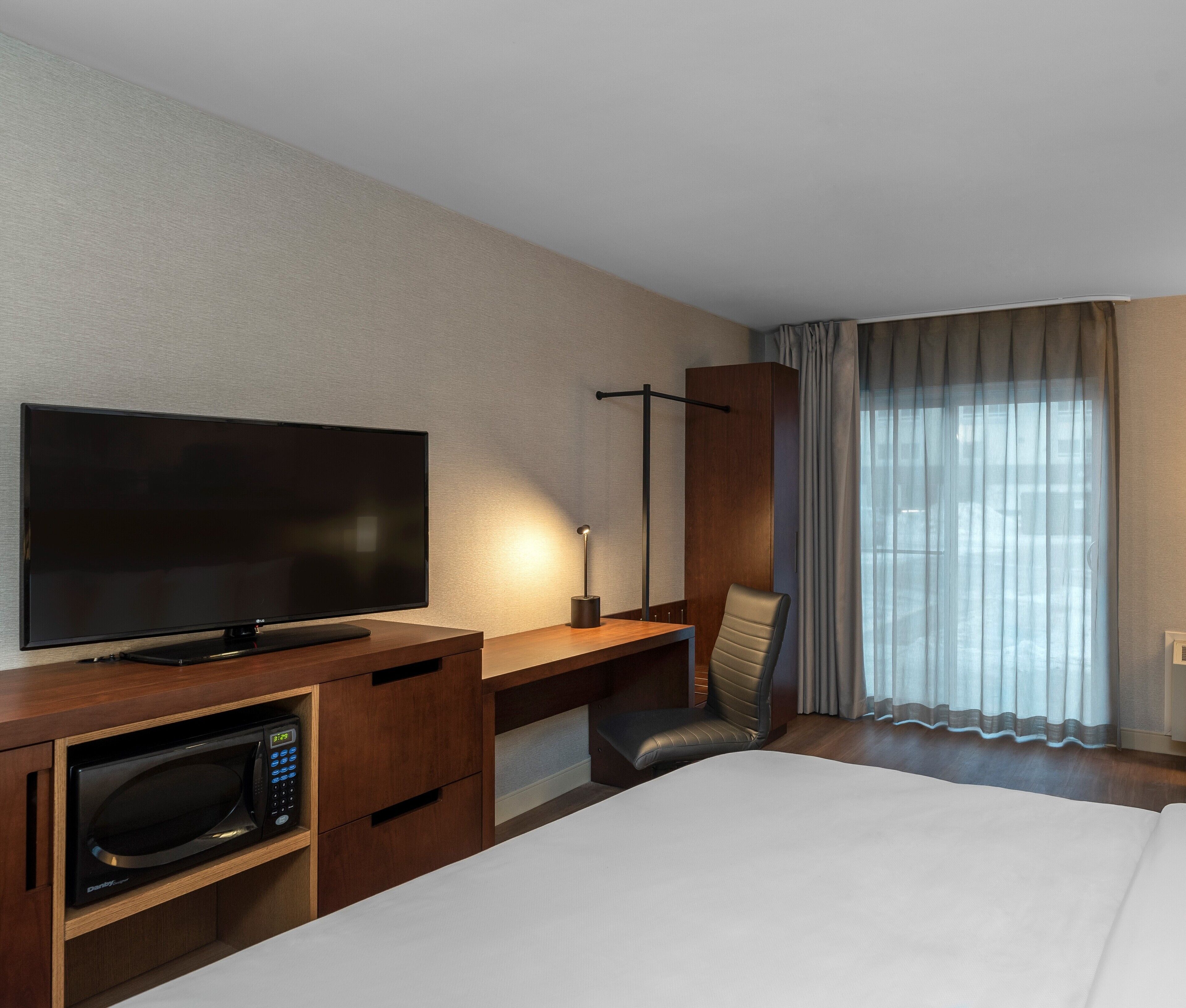 Een hotelkamer met een flatscreen televisie, een magnetron, een bureau, een stoel en een groot raam met gordijnen.