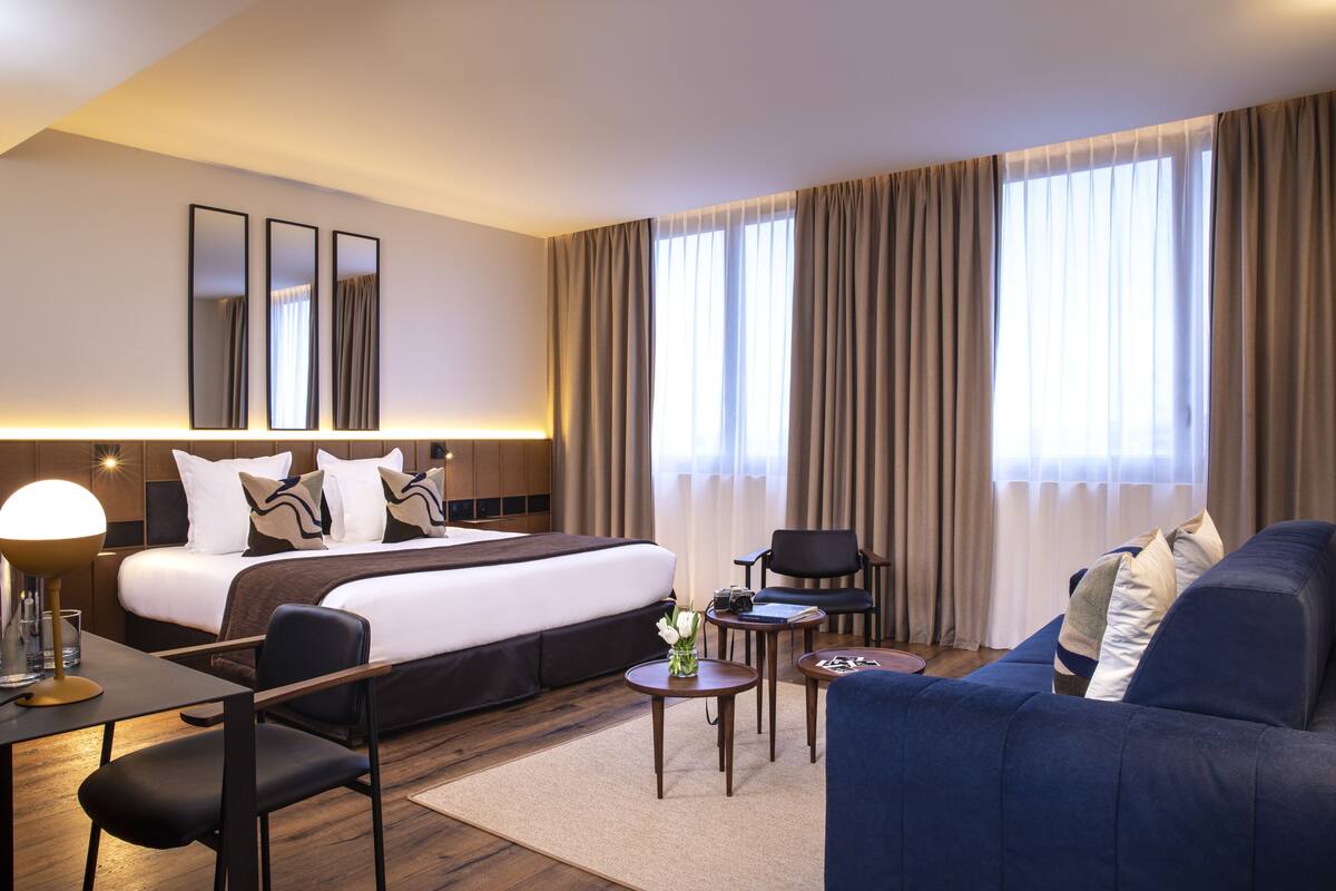 A modern hotel room with a large bed, a sofa, a desk, and a chair. There are two small tables, one with a lamp and a vase of flowers. The room has a large window with curtains.