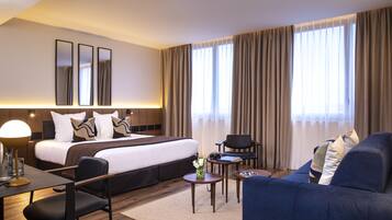 A modern hotel room with a large bed, a sofa, a desk, and a chair. There are two small tables, one with a lamp and a vase of flowers. The room has a large window with curtains.