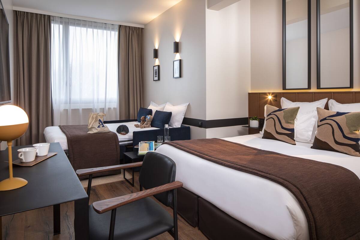 A hotel room with a large bed, a desk with a lamp, a chair, and a small table with cups and a book.