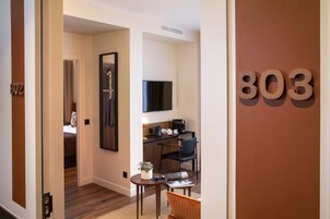 Habitación de hotel con piso de madera, un escritorio con silla, una mesita con planta, una cama con almohada y un televisor montado en la pared.