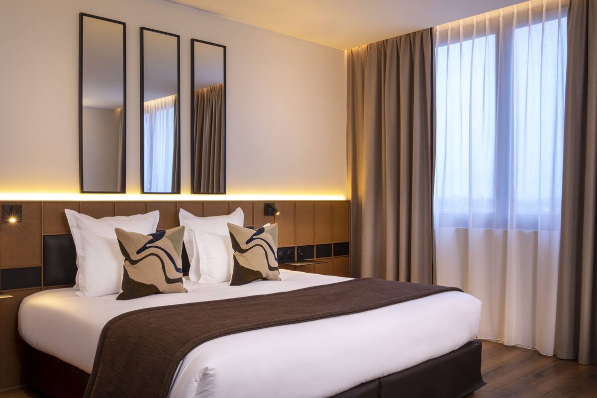 A hotel room with a large bed, a headboard with controls, a wall-mounted mirror, and a window with curtains.