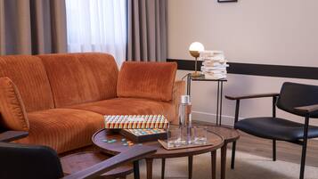 A modern hotel room with a brown sofa, a small round table with a board game, a stack of books, and a lamp.