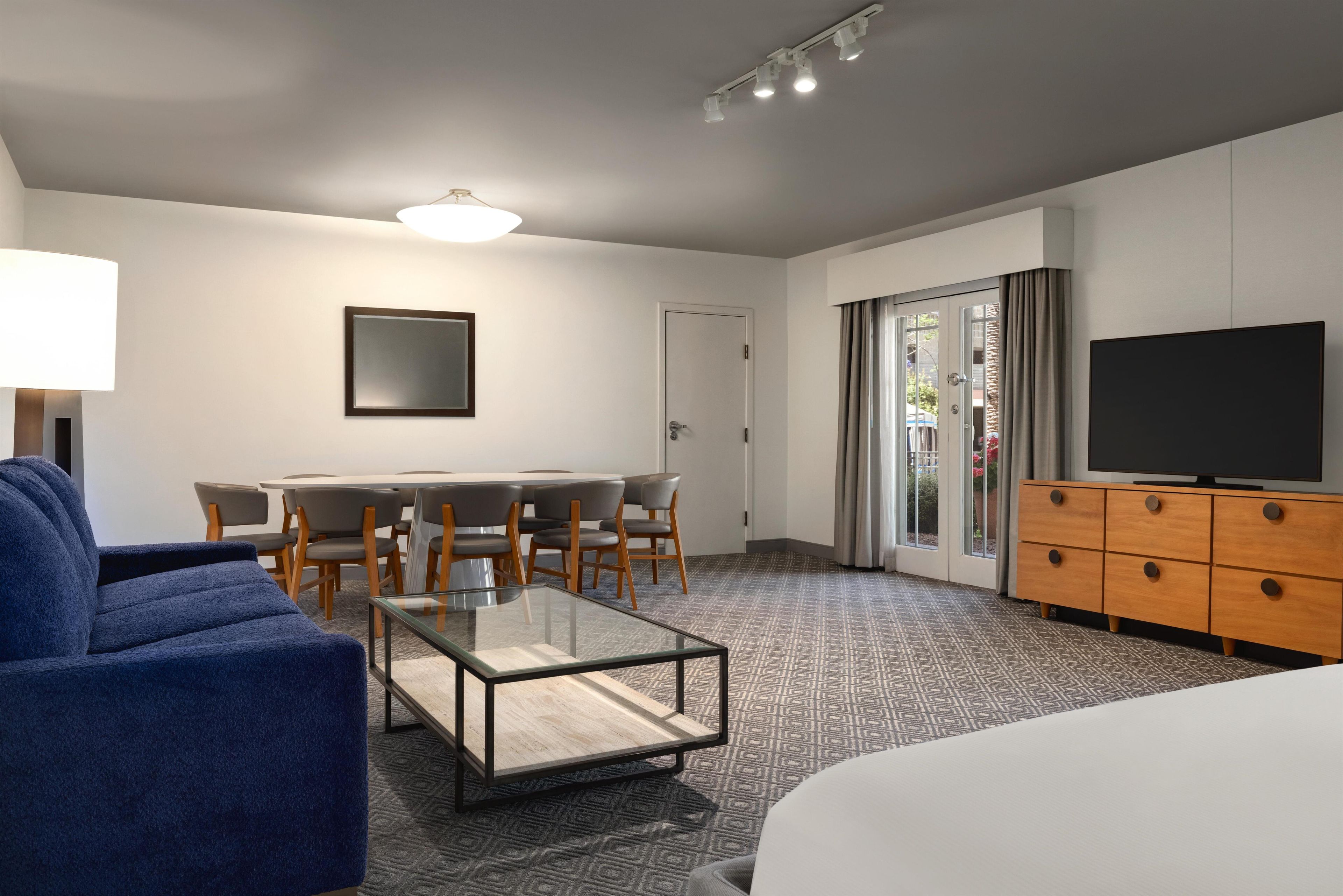 A modern hotel room with a glass coffee table, a sofa, a TV, a dining area with chairs, and a sliding glass door.