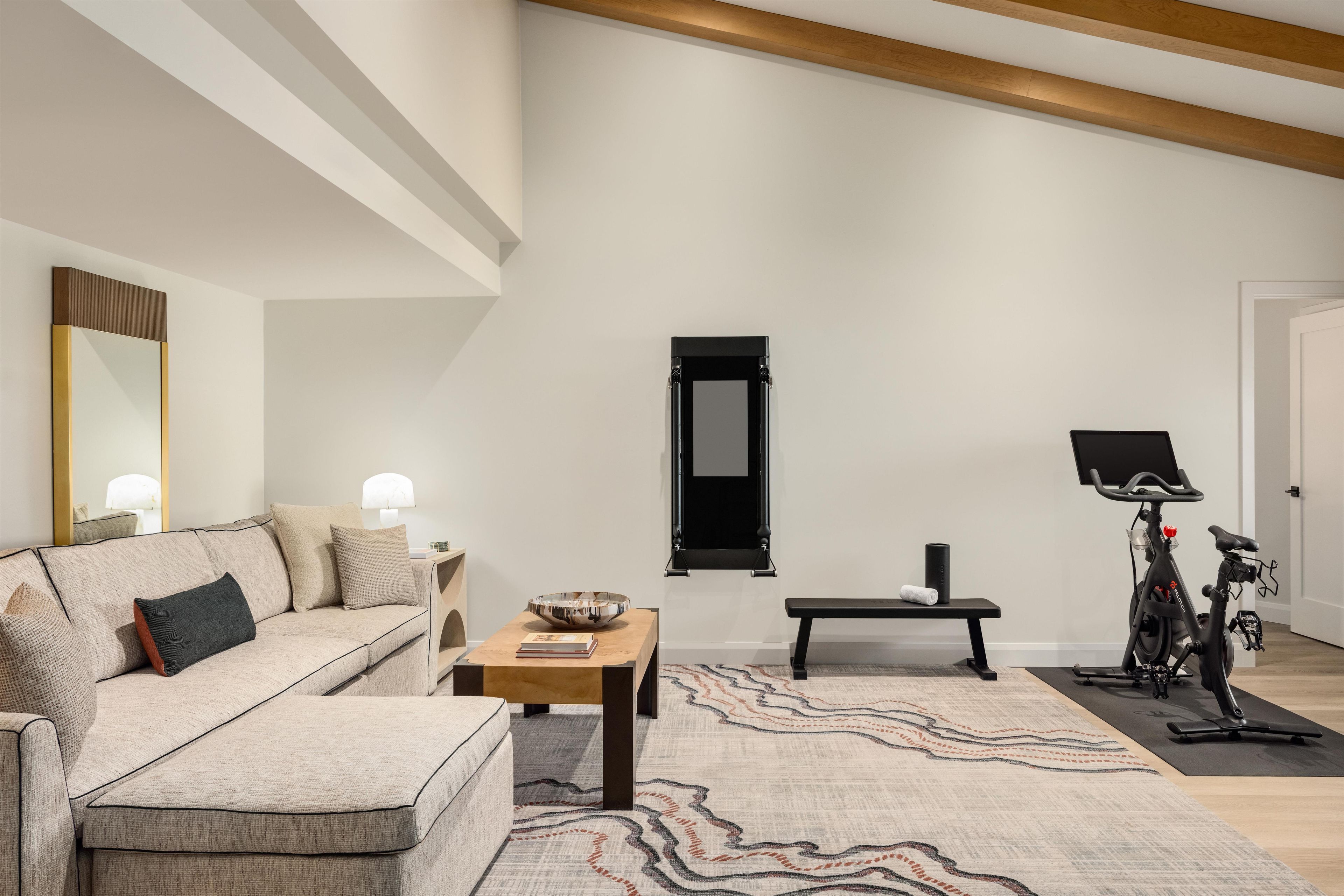 A spacious living room with a high ceiling, a large mirror, a sofa, a coffee table, a rug, and a stationary exercise bike.