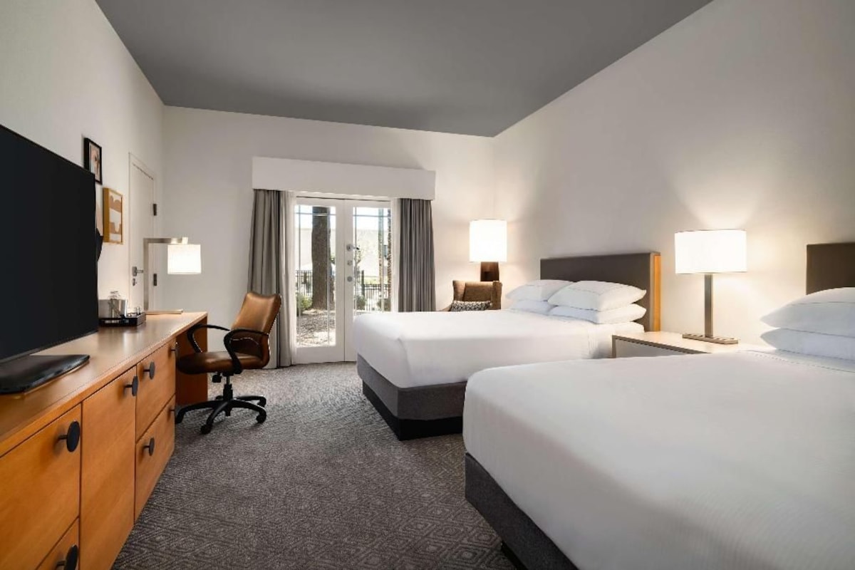 Quarto de hotel com duas camas, uma escrivaninha com televisão, uma cadeira e vista através de uma porta de vidro.