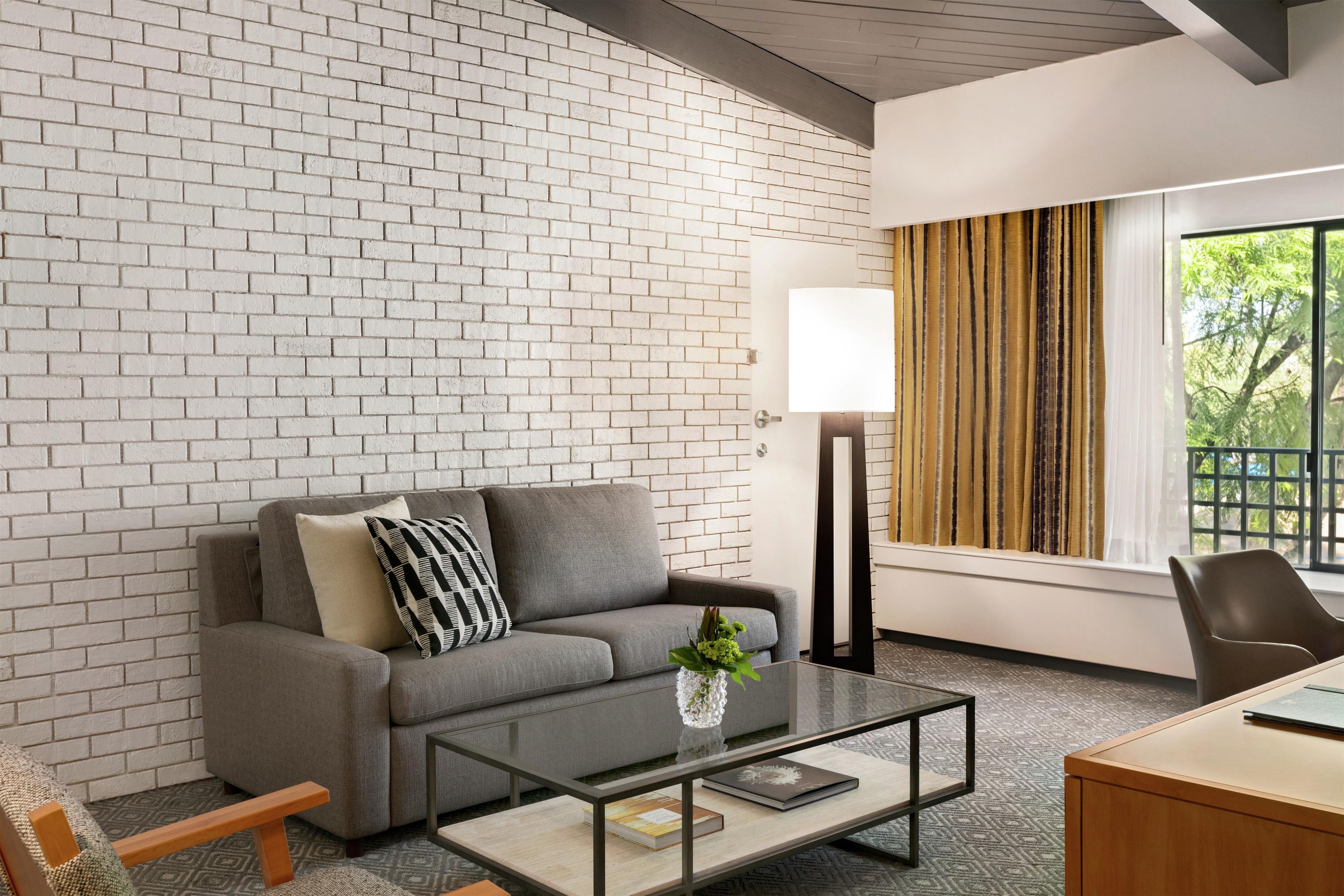 A modern room with a brick wall, a grey sofa, a glass coffee table, a desk, and a lamp.