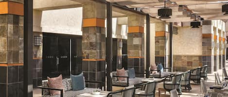 An outdoor dining area with stone columns, cushioned seating, and tables set for dining.