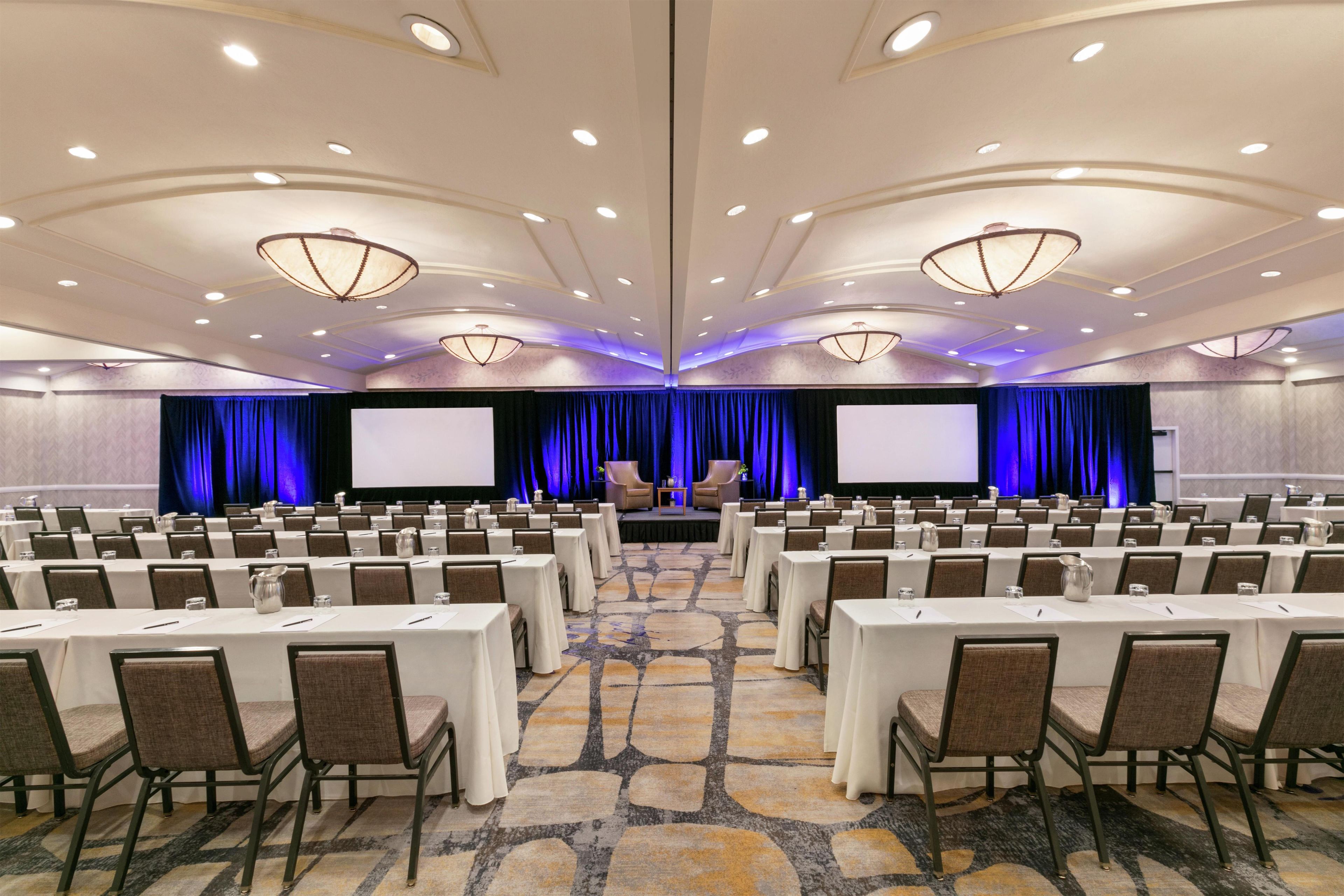 A conference room with rows of tables and chairs, a stage with a screen, and a carpeted floor.
