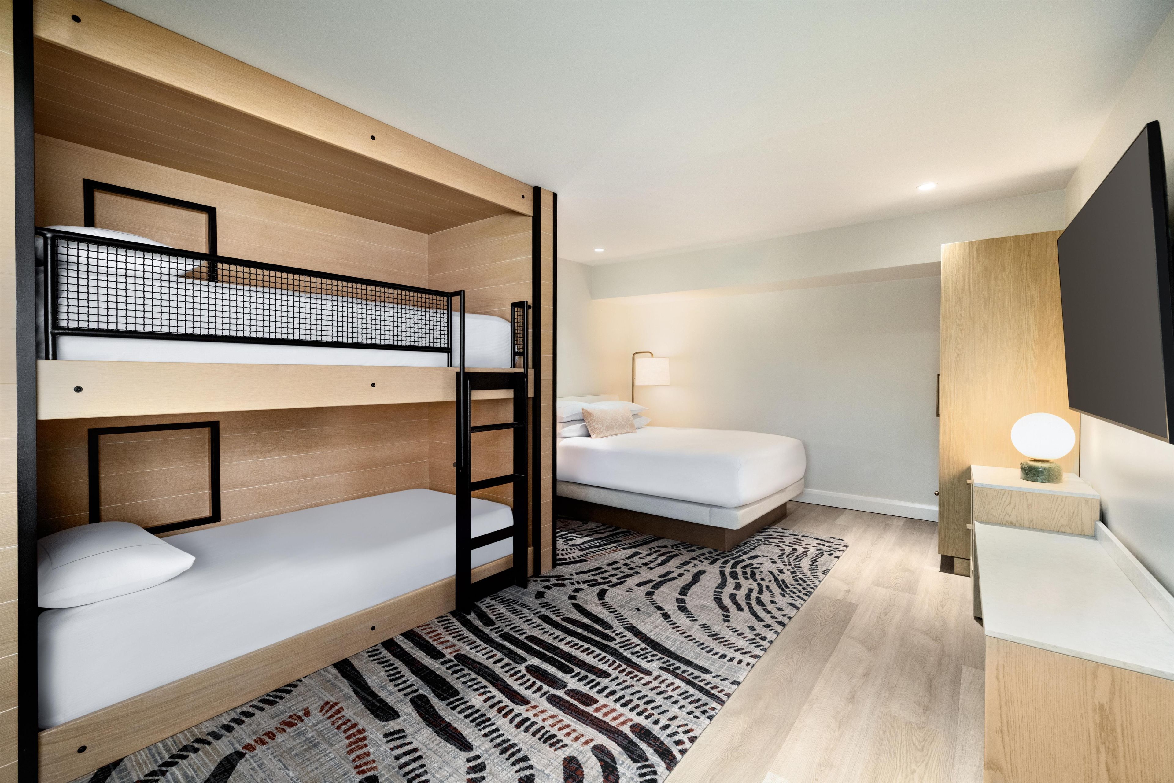 A modern hotel room with bunk beds, a single bed, a bedside table with a lamp, and a patterned rug.