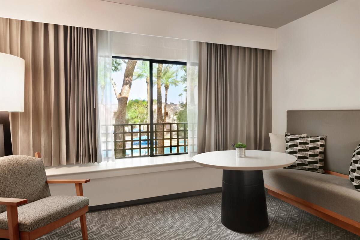 A hotel room with a sofa, a round table, a chair, a window with curtains, and a view of palm trees.