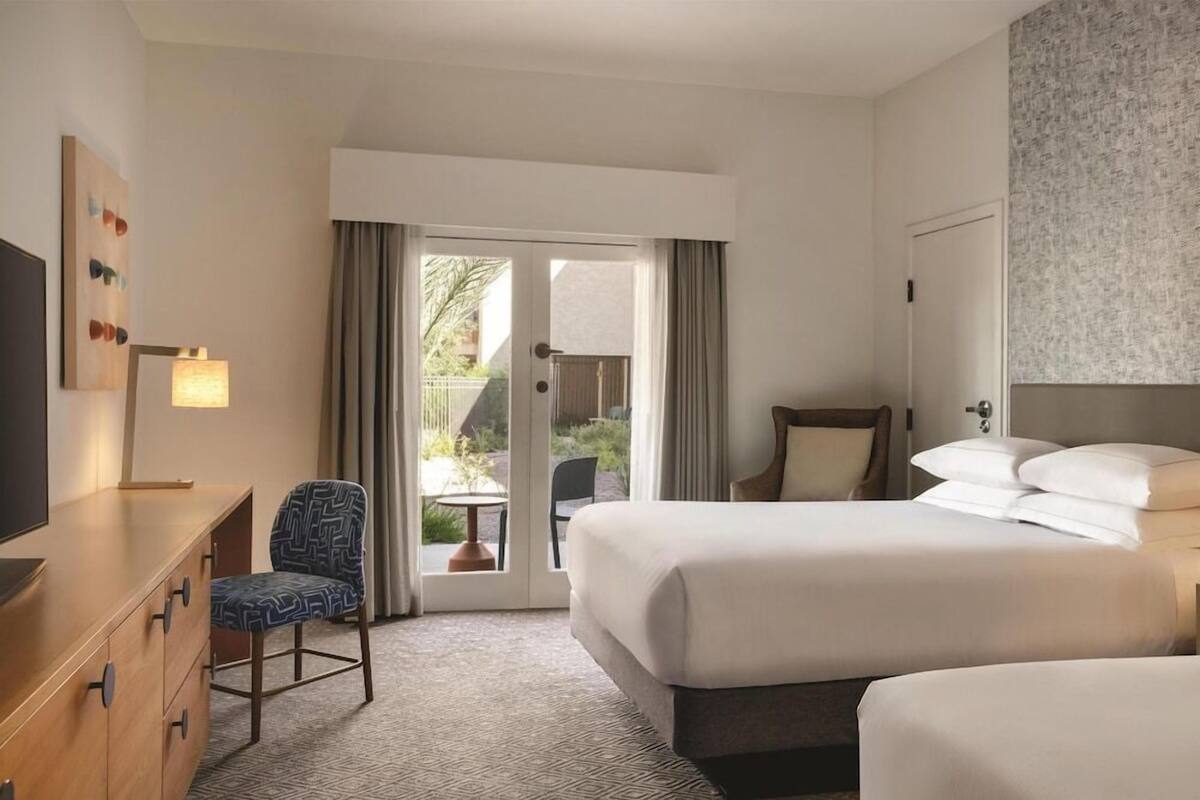 A hotel room with two beds, a desk, a chair, a TV, and a sliding door to a balcony.