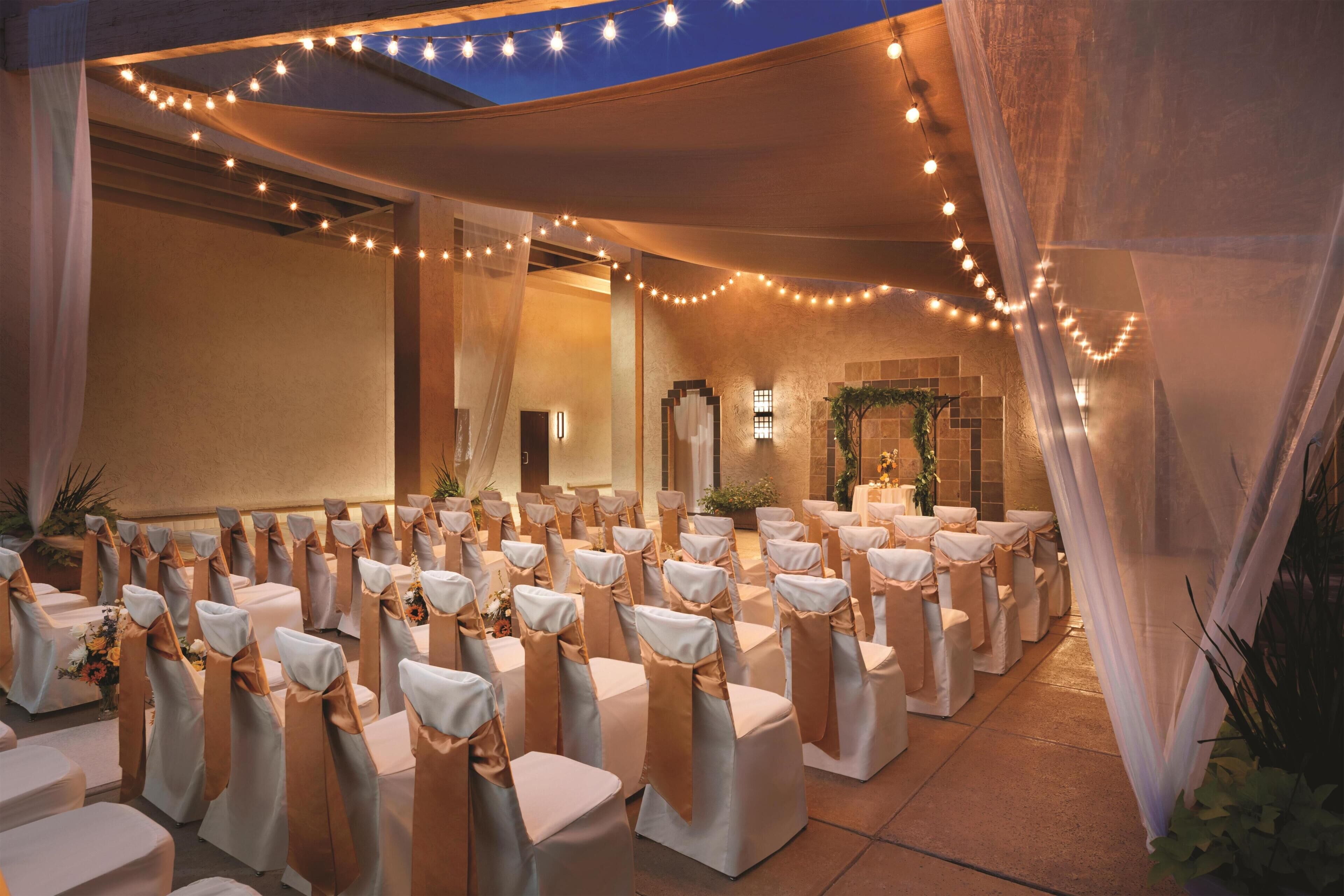 A banquet hall with rows of tables, chairs with white and gold sashes, string lights, and a large window.
