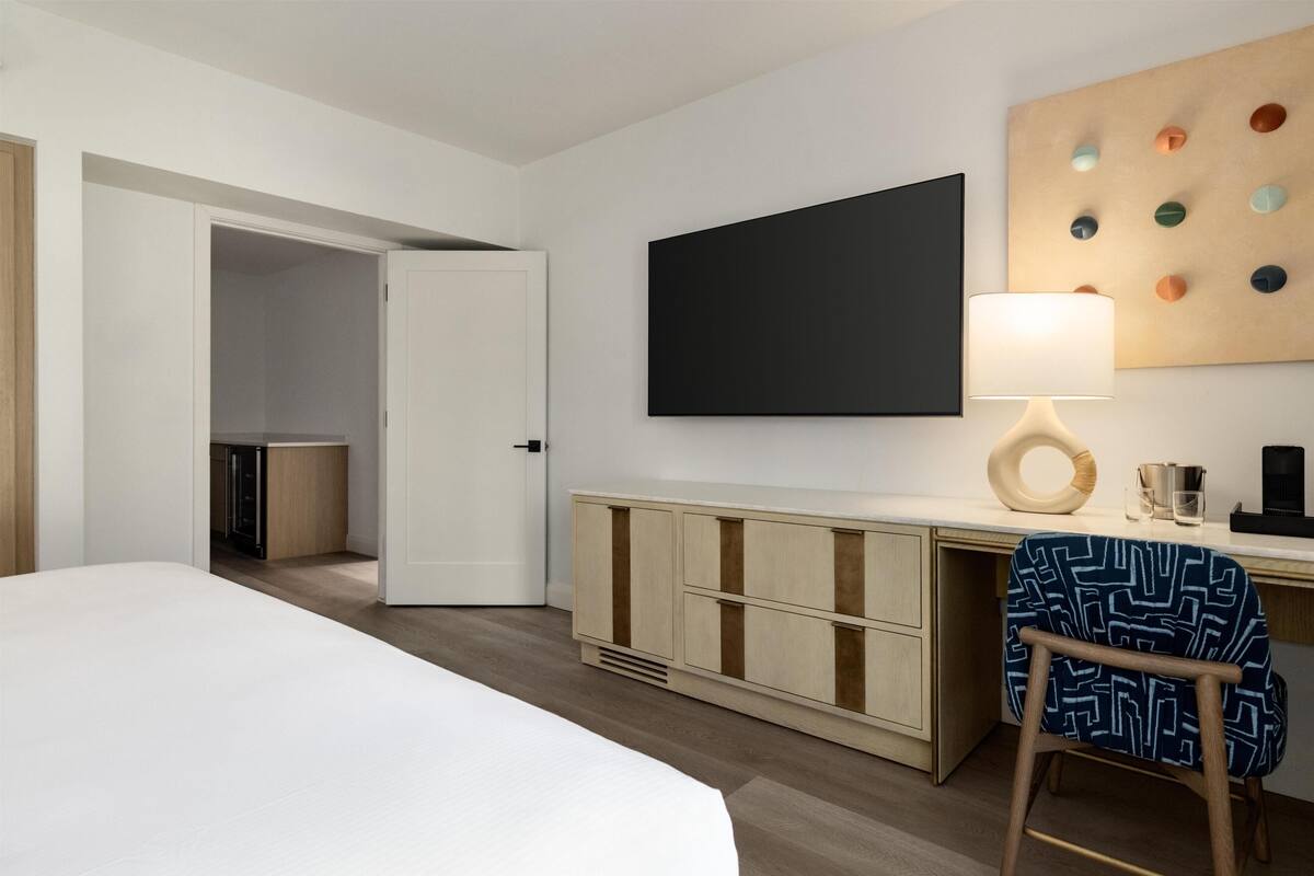A hotel room with a bed, a desk with a lamp, a wardrobe, and a wall-mounted TV.