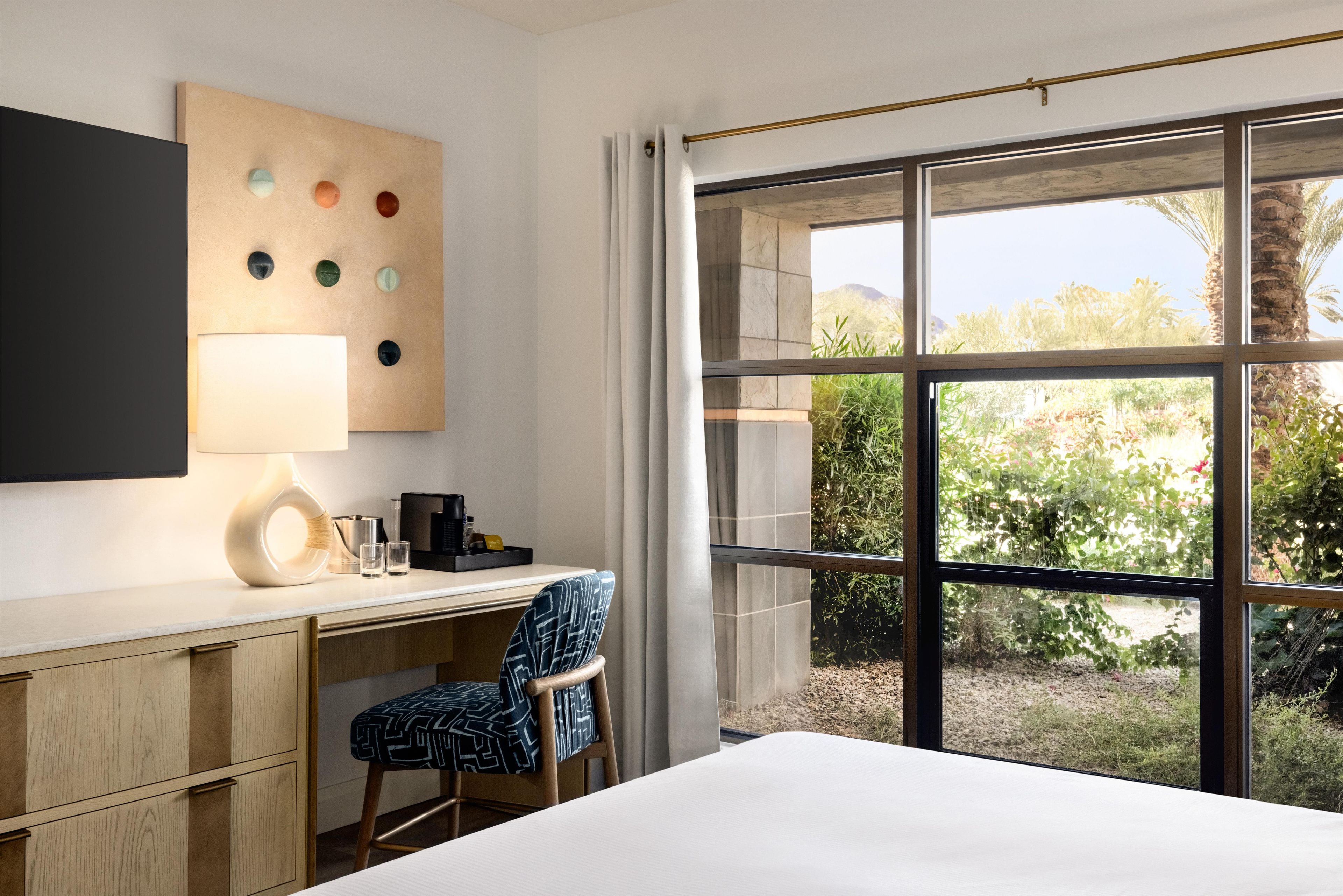 A hotel room with a desk, chair, bed, and a large window offering a view of greenery and a mountain.