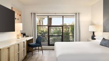 A hotel room with a bed, a desk, a chair, a TV, and a window with a view of trees and buildings.