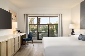 Habitación de hotel con cama, escritorio, silla, televisión y ventana con vistas a árboles y edificios.