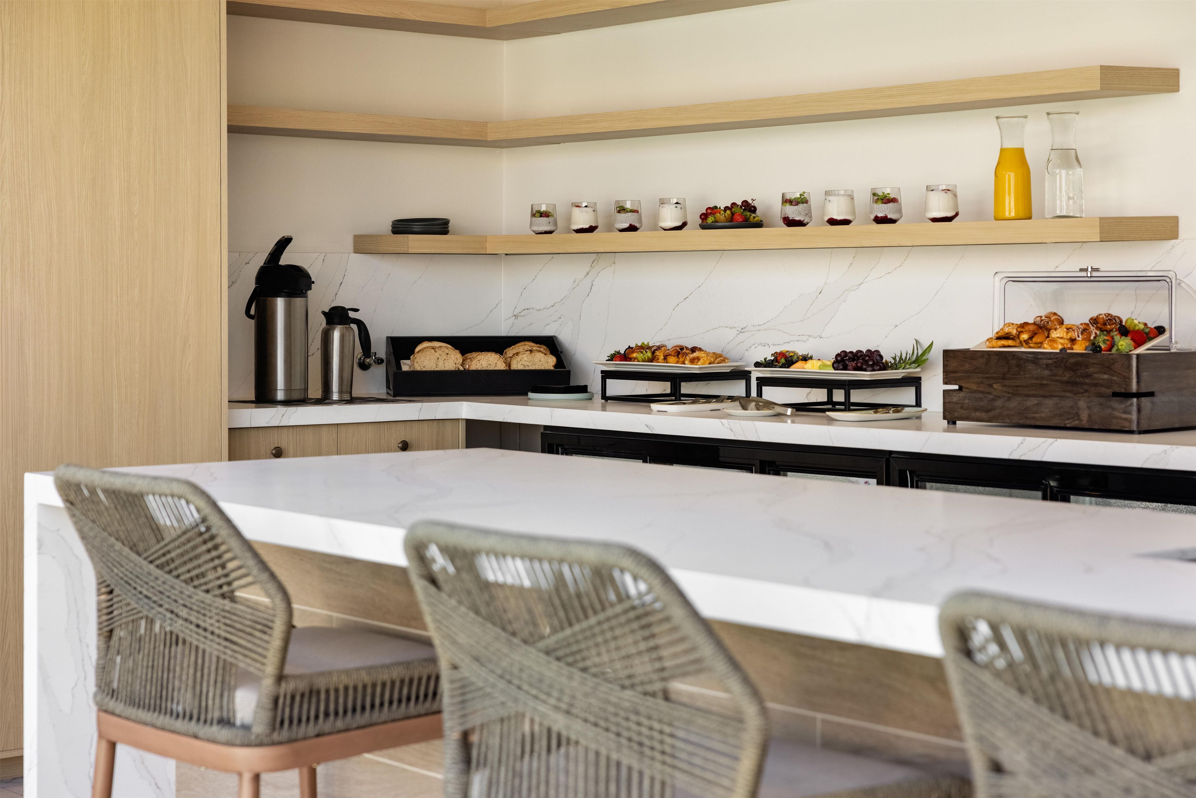 A modern kitchen with a marble countertop, wicker chairs, and a variety of food items displayed on shelves.