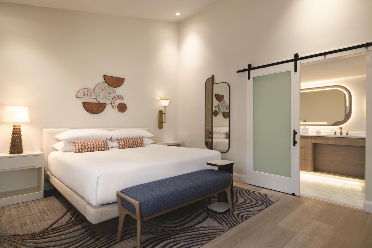 A modern hotel room with a large bed, a bench, a wall-mounted mirror, a bedside table with a lamp, and a sliding door to the bathroom.