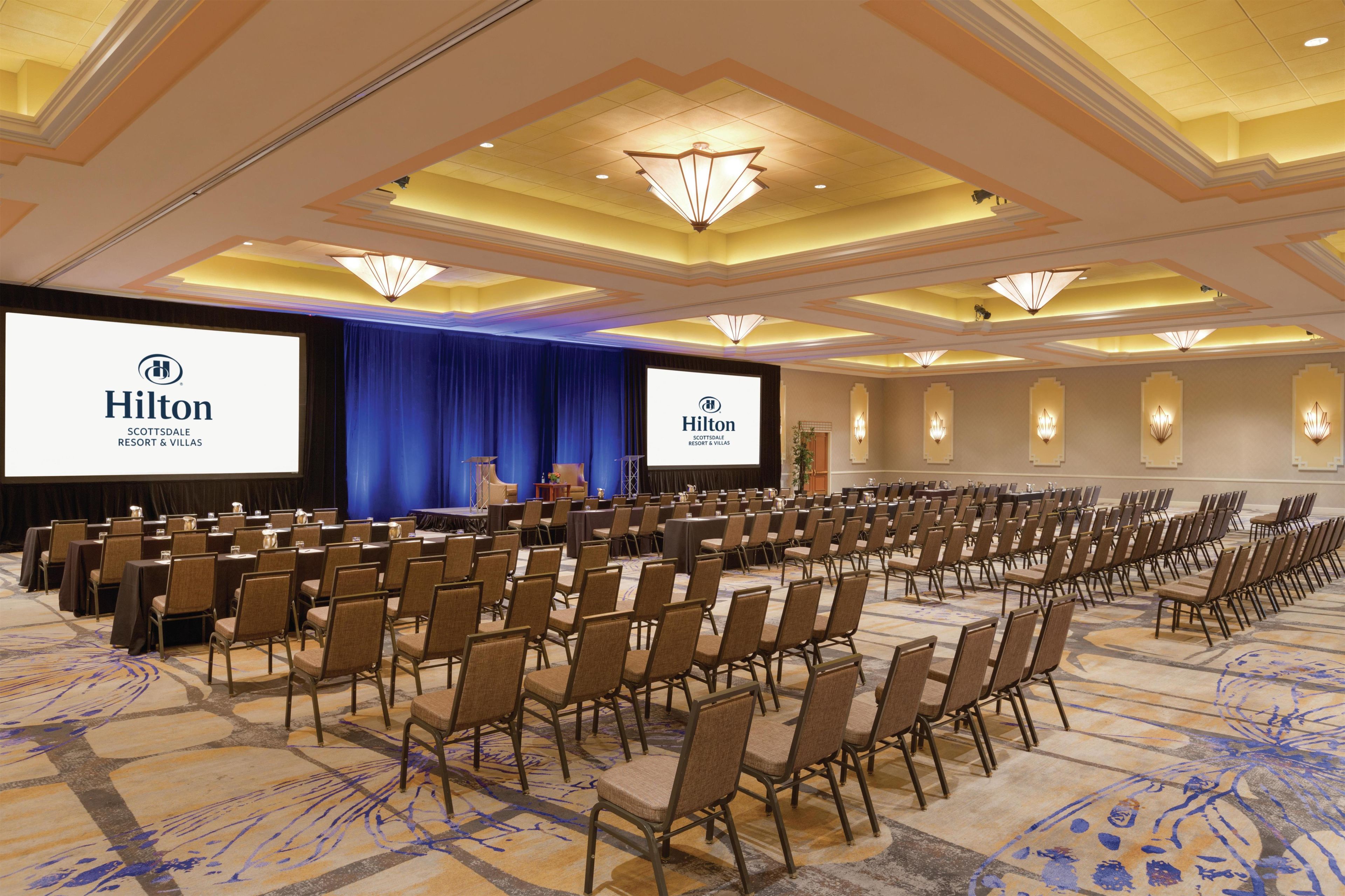 Une salle de conférence avec des rangées de chaises, une scène sur laquelle est installé un écran affichant le logo de Hilton, et un sol recouvert de moquette.