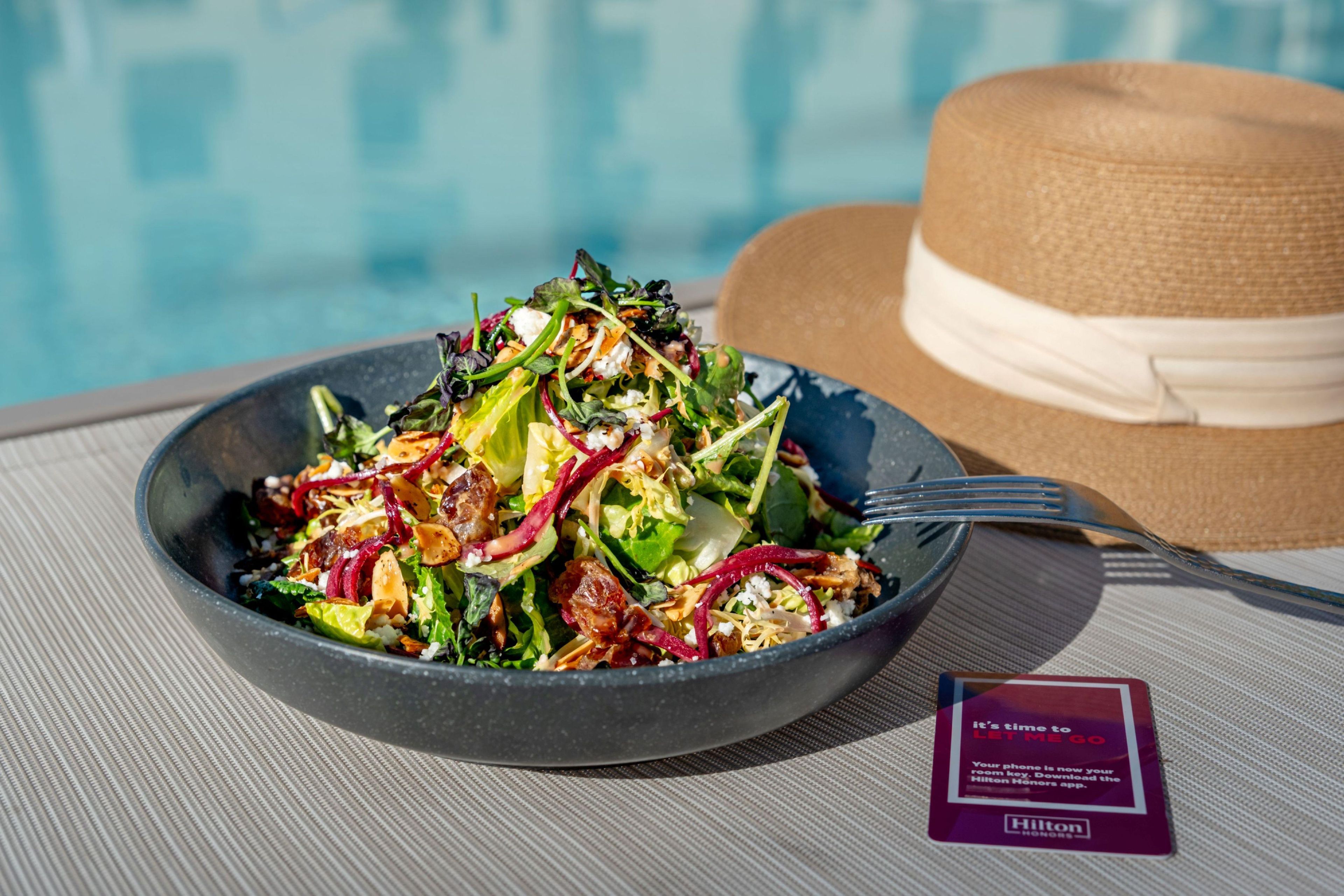 A bowl of salad with a fork on a table near a pool, a straw hat, and a sign with text.