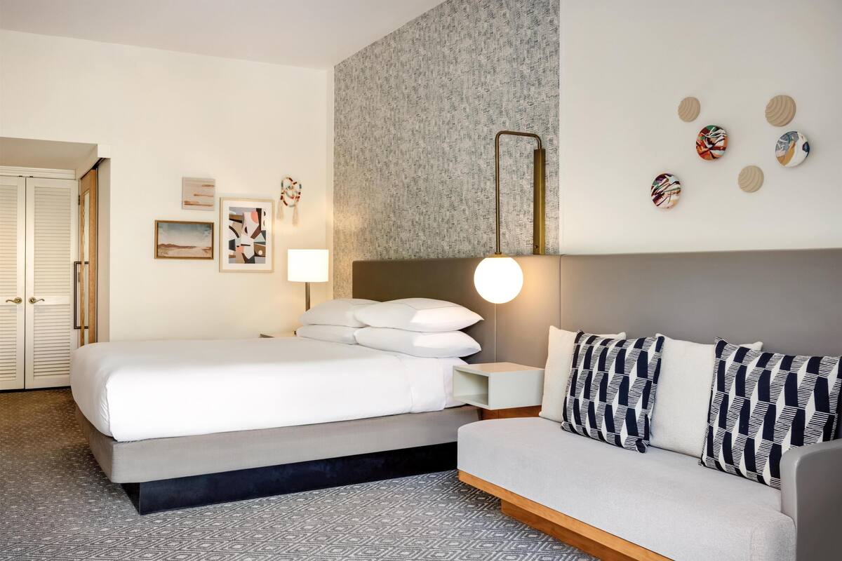 A hotel room with a bed, a sofa, a wall-mounted lamp, and framed artwork.