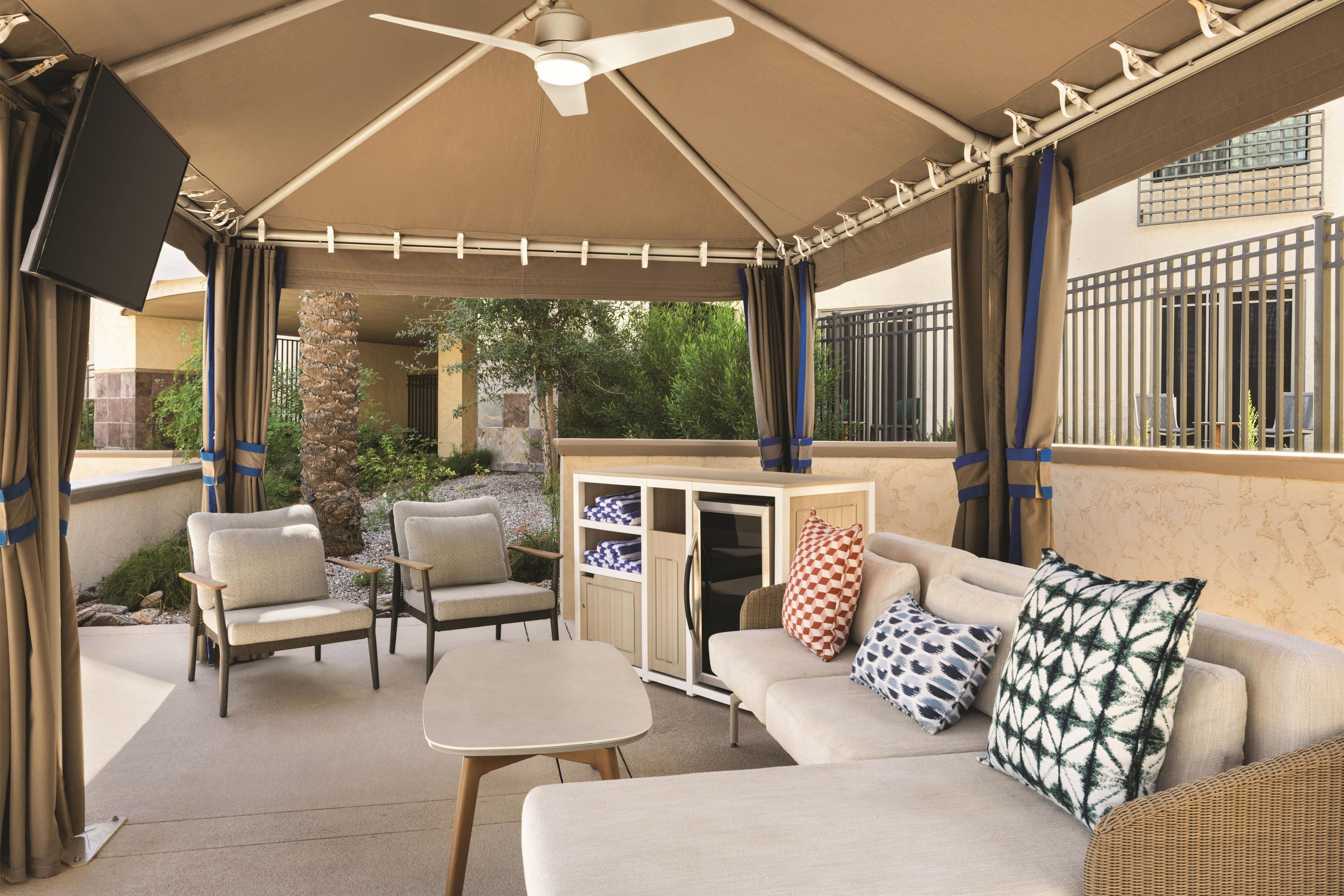 An outdoor patio with a ceiling fan, cushioned seating, a small table, and a cabinet with storage.