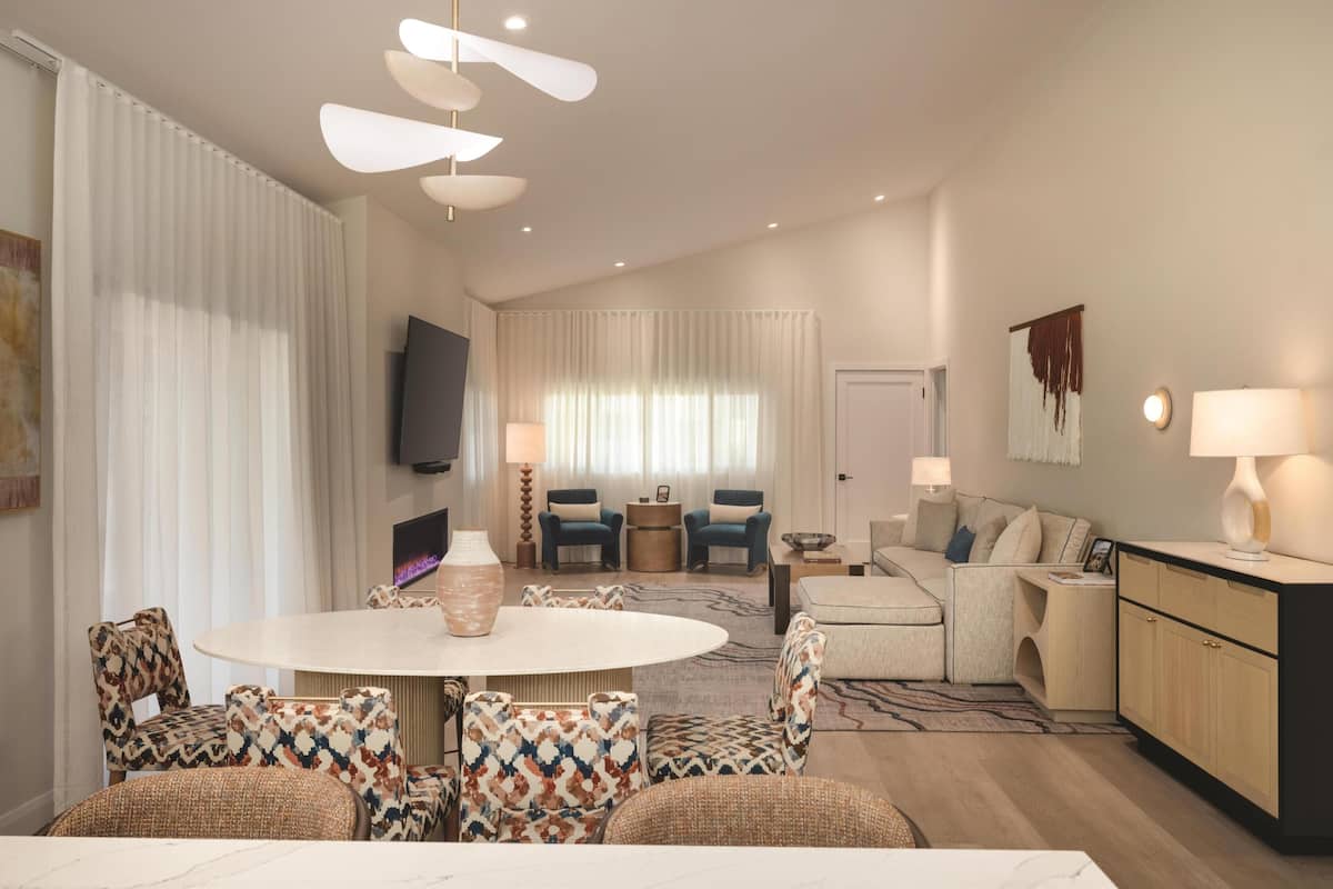 A modern living room with a round dining table, patterned chairs, a sofa, a TV, and a kitchen area with cabinets and a countertop.