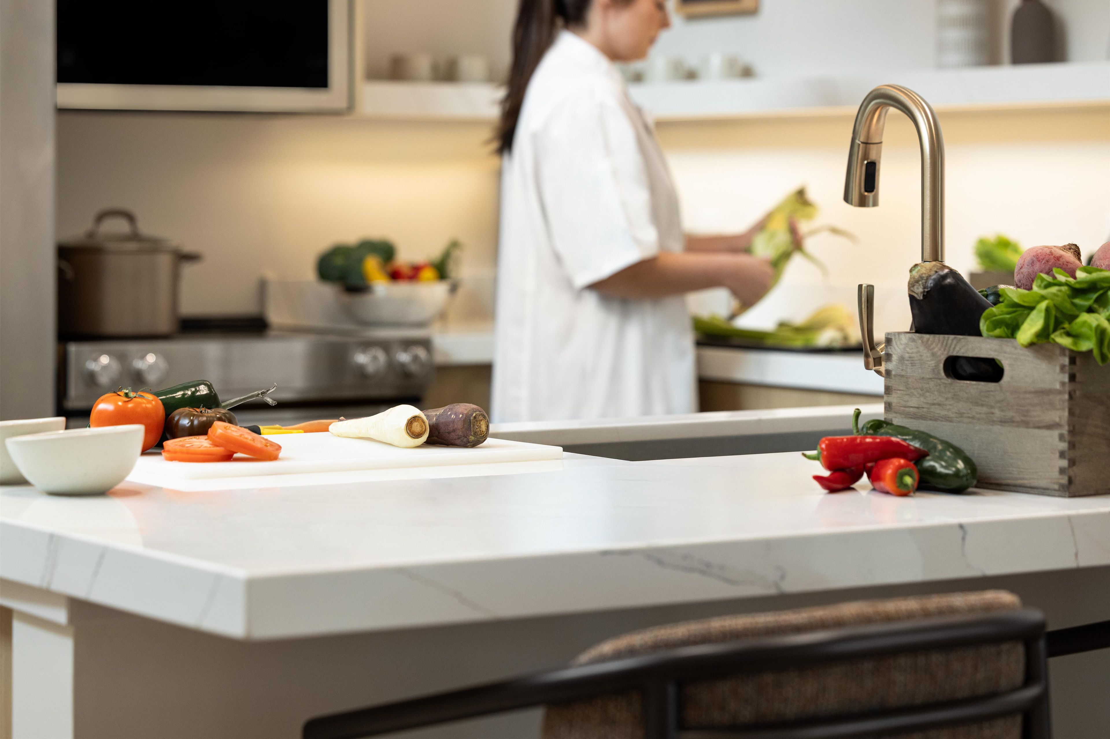 A kitchen with a countertop, a cutting board, vegetables, and a person preparing food.