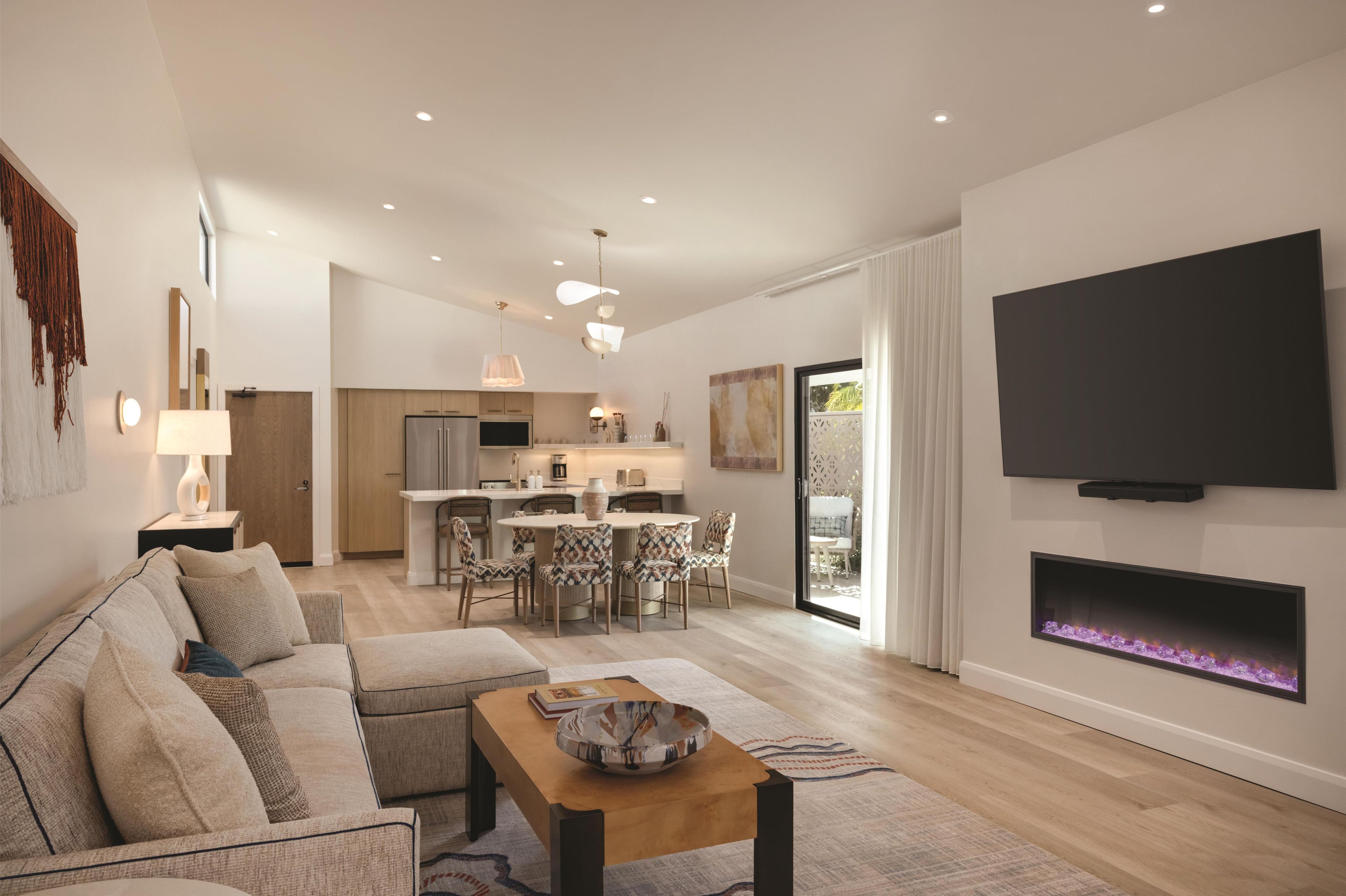 Un salón moderno con sofá, mesa de centro, chimenea, televisión y una cocina al fondo.