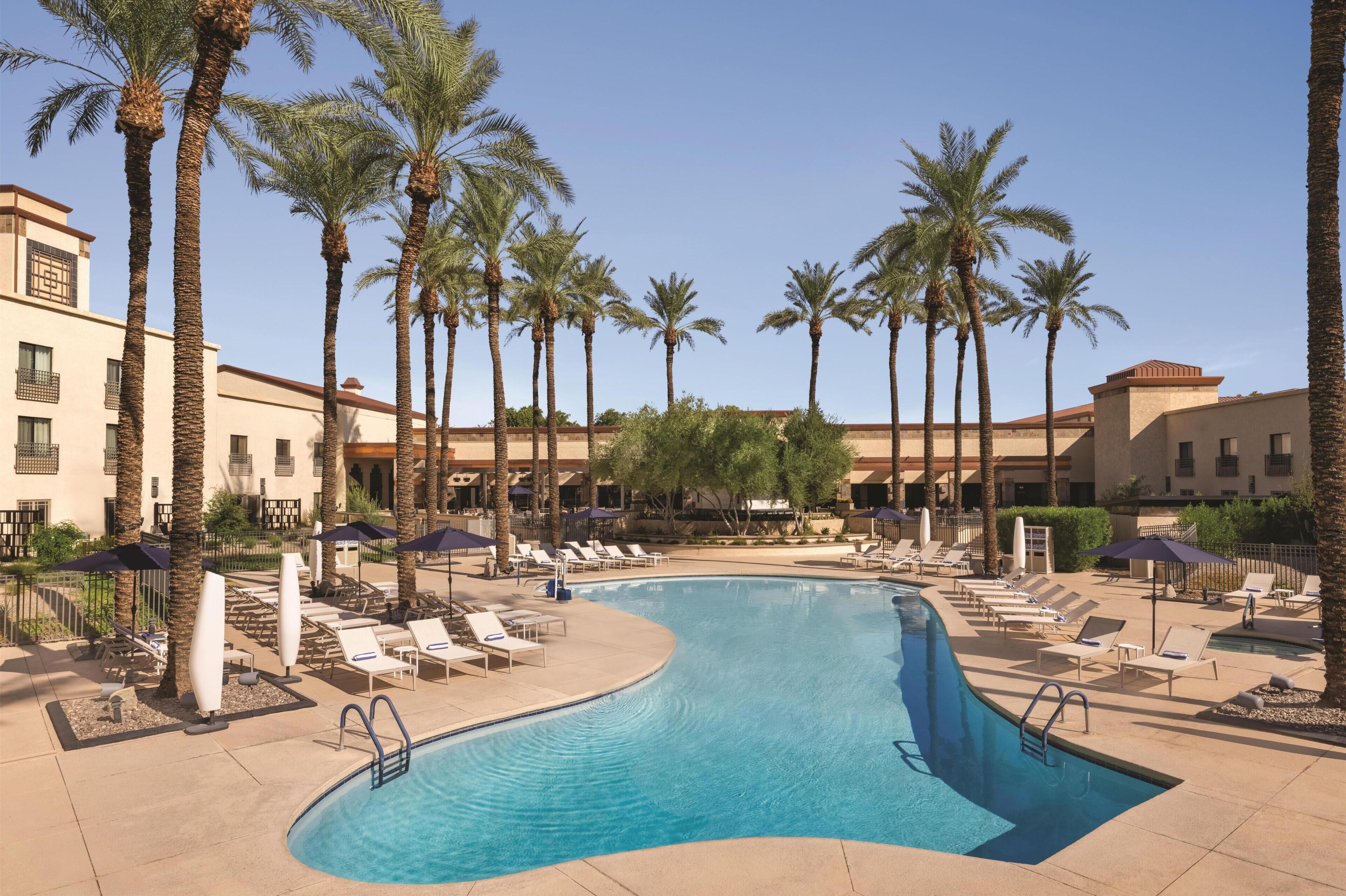 Ein Swimmingpool mit Liegestühlen und Palmen, umgeben von Hotelgebäuden.