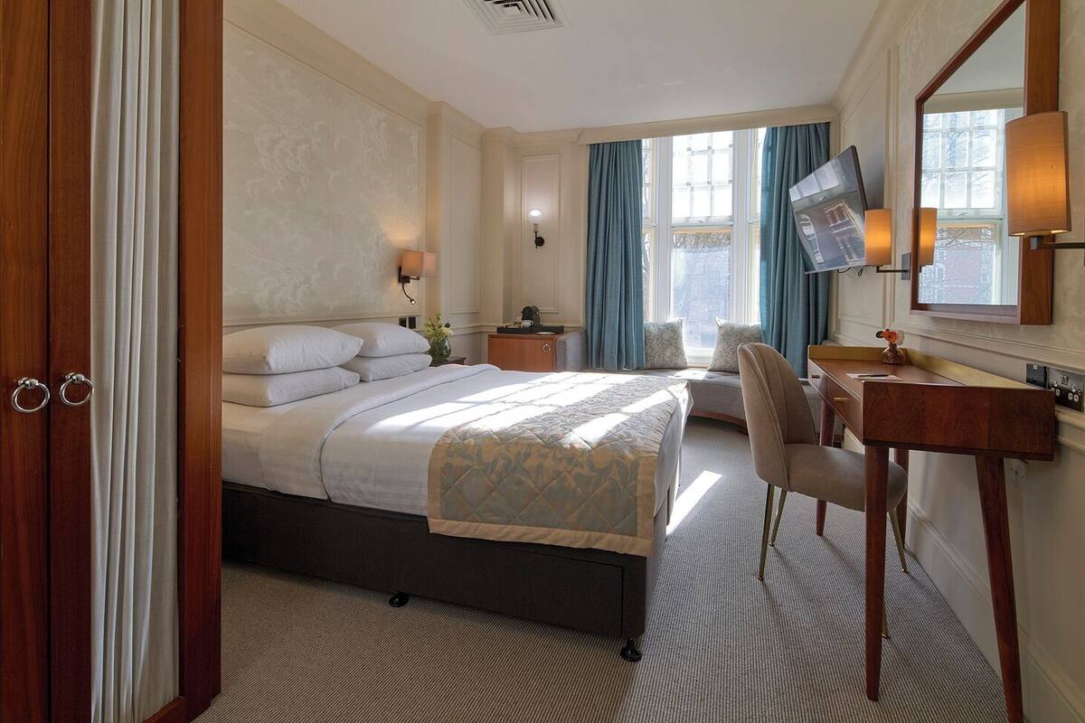 A hotel room with a large bed, a desk with a chair, a vanity with a mirror, and a television on the wall.
