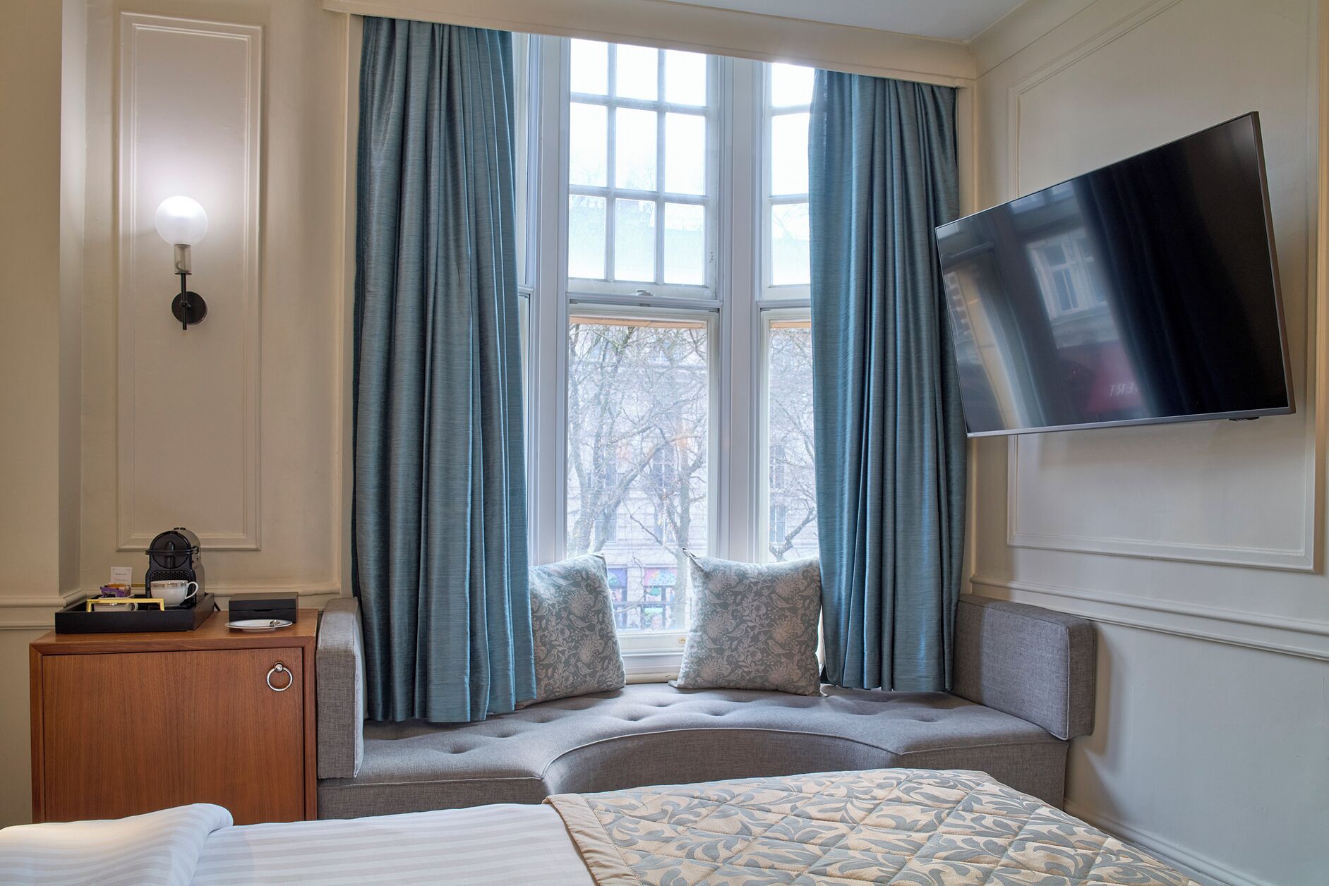 A hotel room with a large window, a flat-screen TV mounted on the wall, a bed with a patterned bedspread, and a small wooden dresser with a lamp and a tray of items.