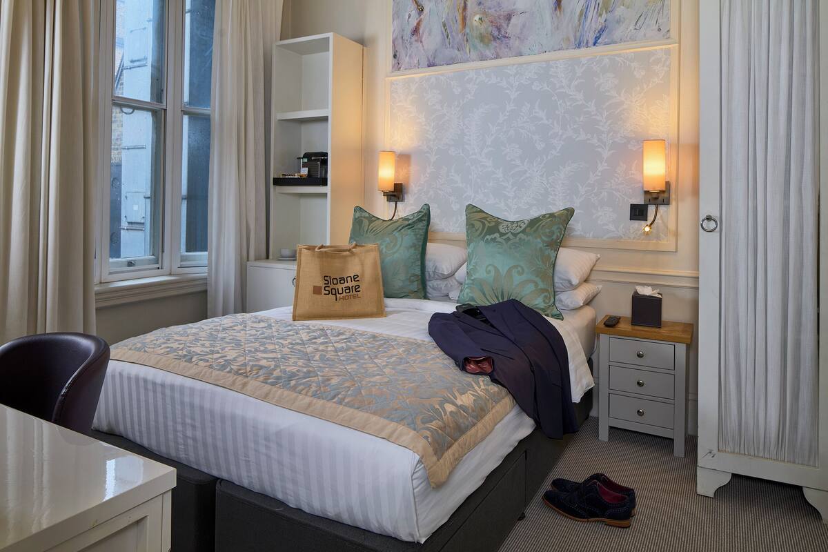 A hotel room with a bed, bedside tables, a desk, and a chair. There is a window with curtains, a painting on the wall, and a bag on the bed.