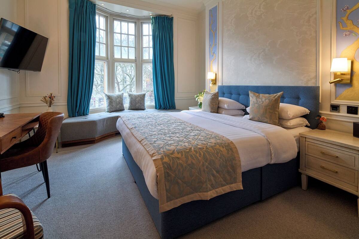 A hotel room with a large bed, a desk, a chair, a TV, and a window with curtains.