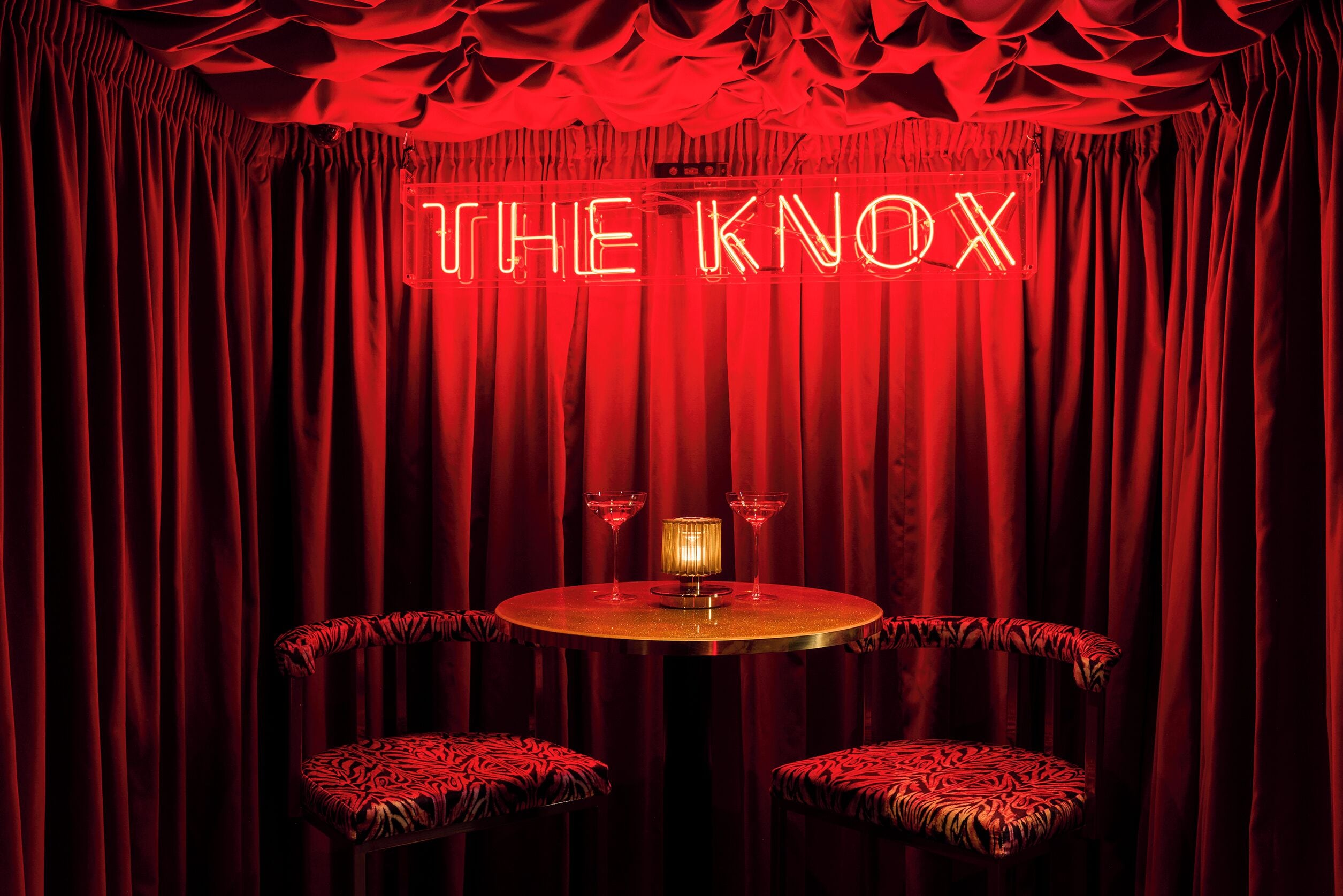 A red-lit room with a neon sign reading "THE KNOX," a round table, two chairs, and two martini glasses.