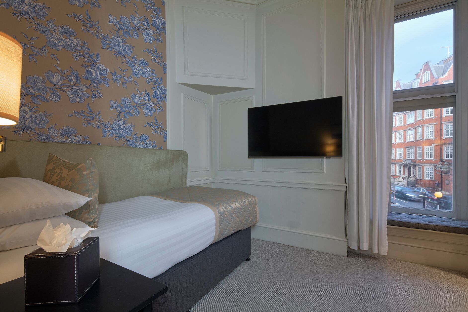 A hotel room with a bed, a bedside table with a tissue box, a flat-screen TV mounted on the wall, and a window with a view of buildings.