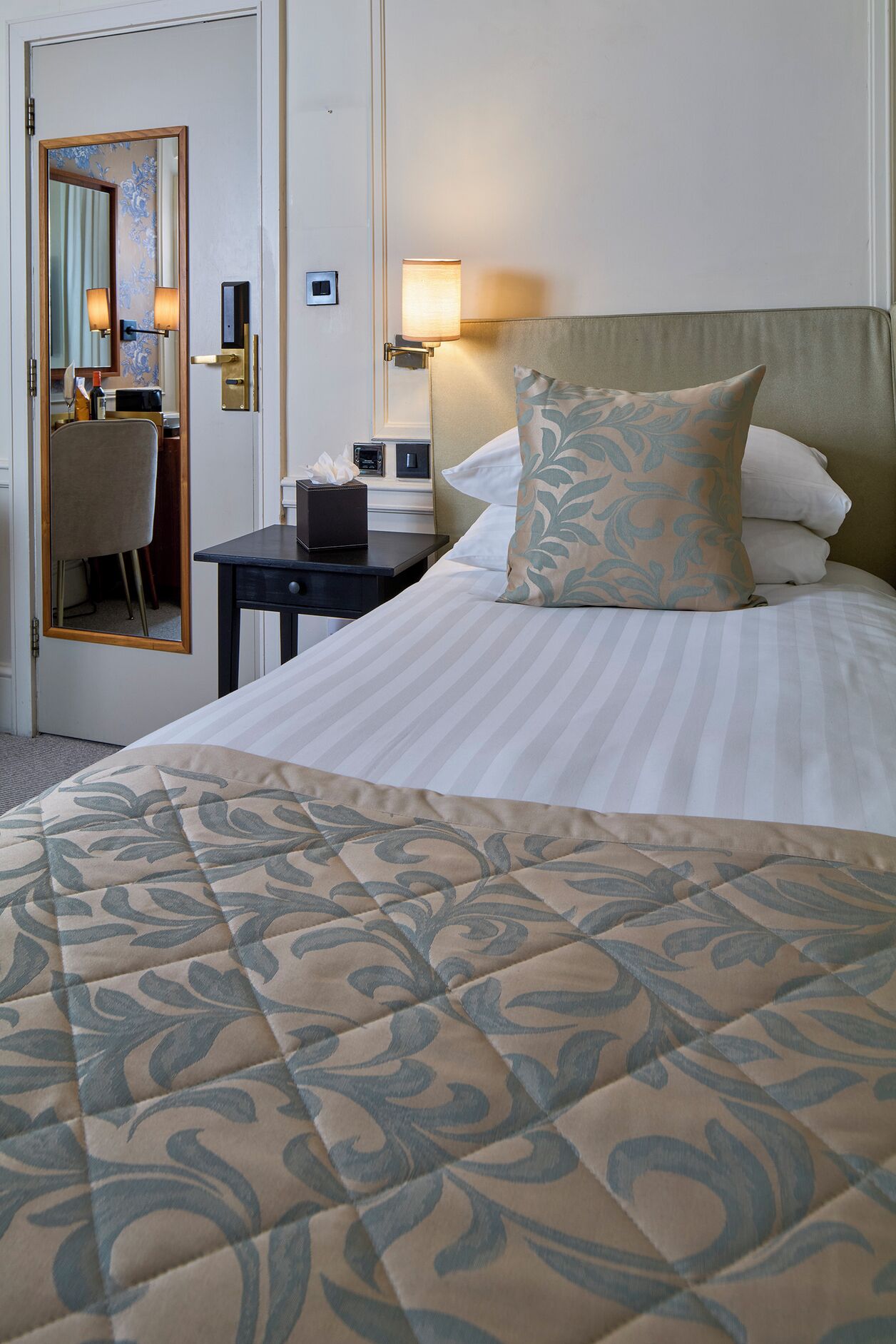 A hotel room with a bed, a bedside table with a lamp, a chair, and a mirror.