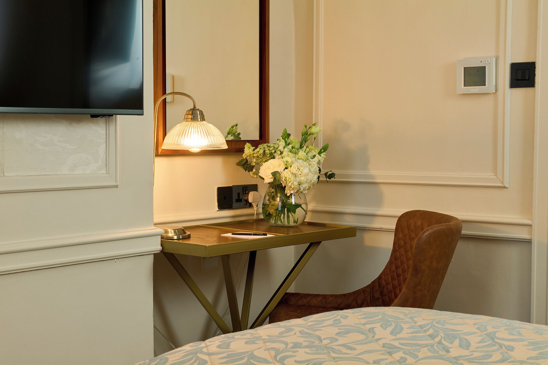 Une chambre d’hôtel comprenant un lit, un bureau avec une lampe, une chaise, un téléviseur et un thermostat fixé au mur.