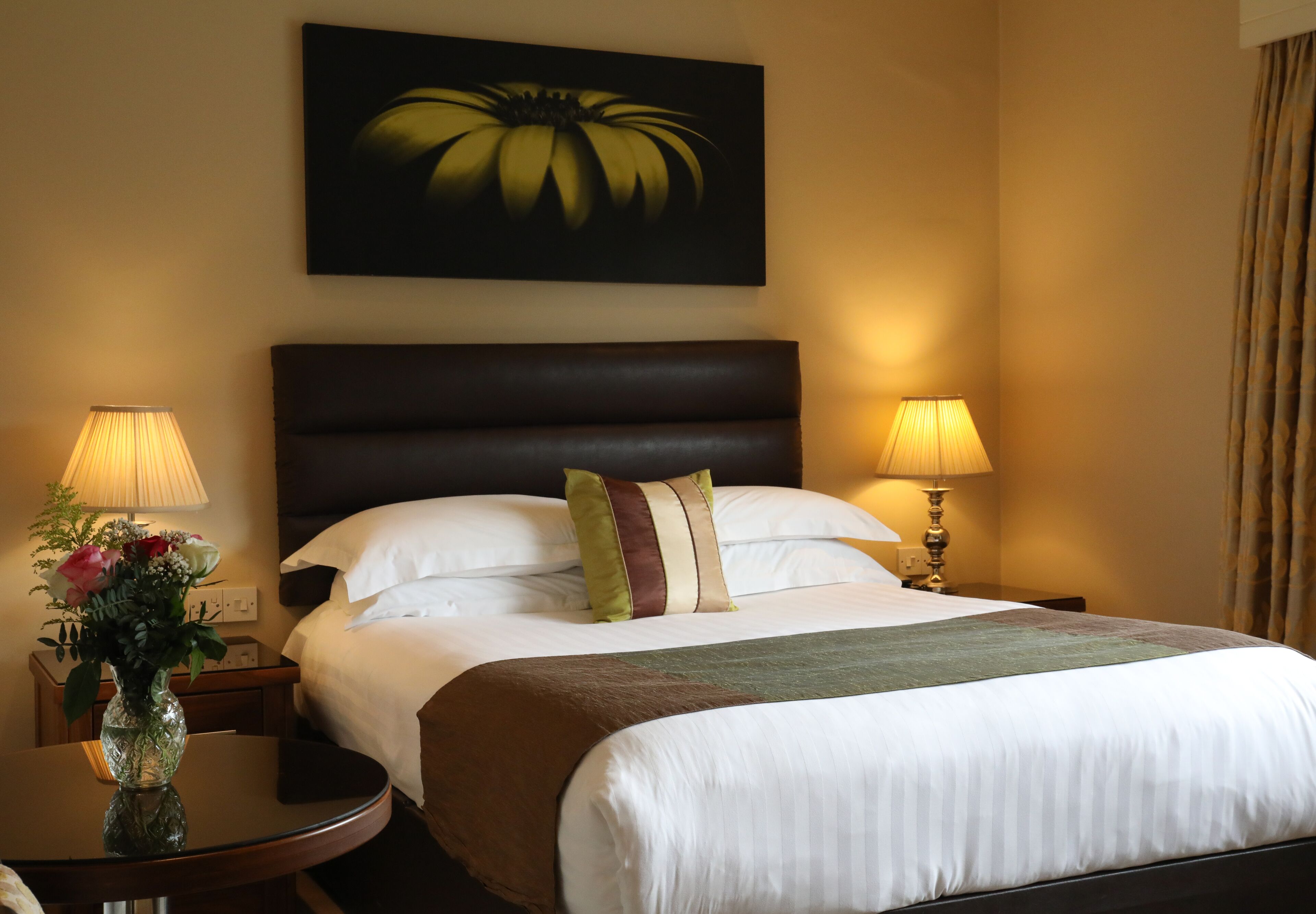 Habitación de hotel con una cama grande, dos lámparas de noche, un jarrón con flores y un cuadro en la pared.