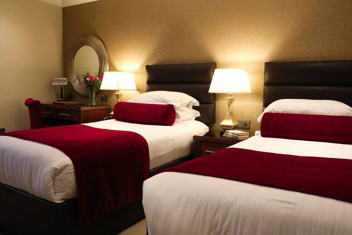 A hotel room with two beds, each with white linens and red throw pillows, flanked by nightstands with lamps and a mirror.