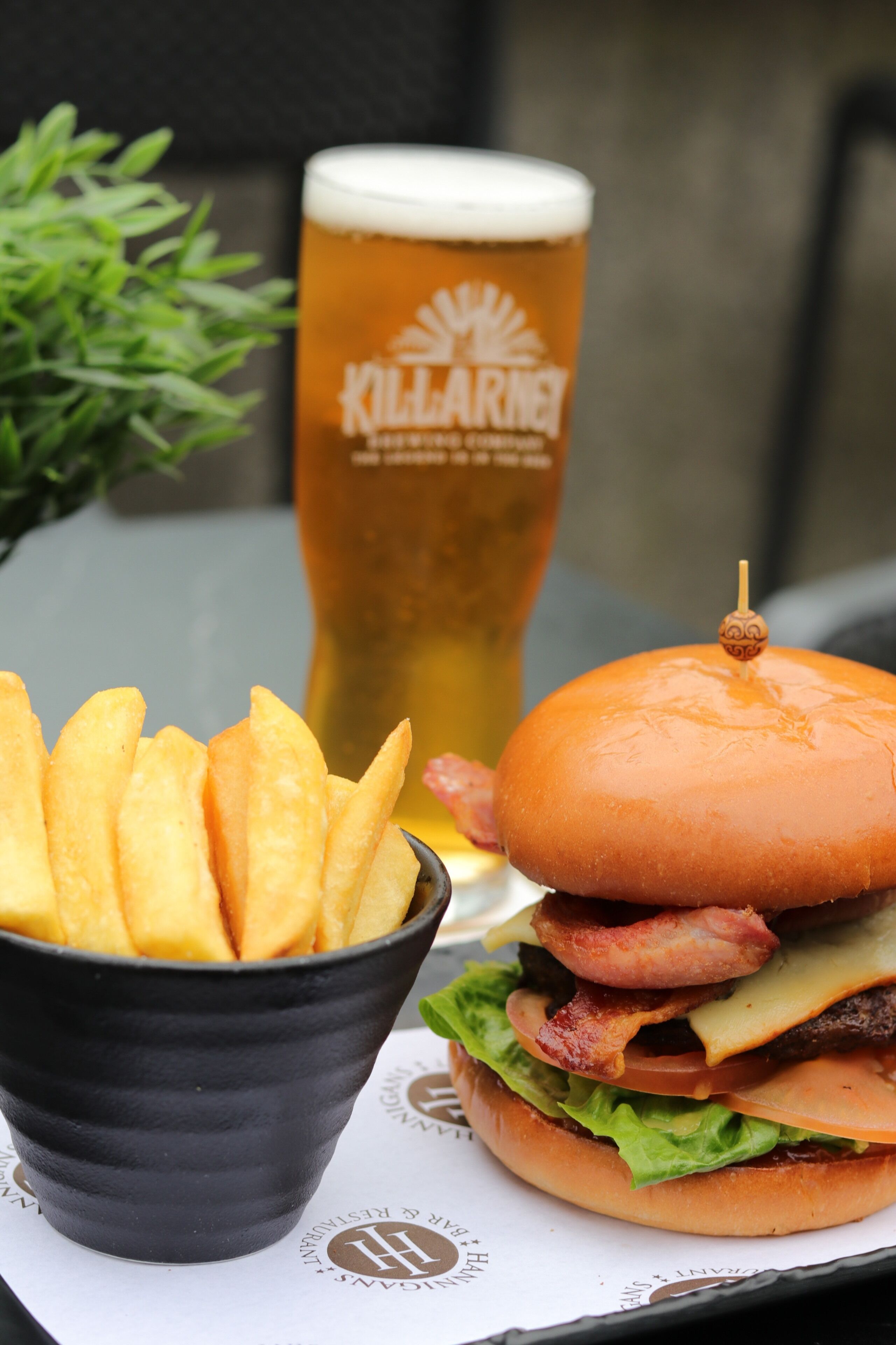 A burger with bacon, cheese, and lettuce, a glass of beer, and a bowl of fries on a table.