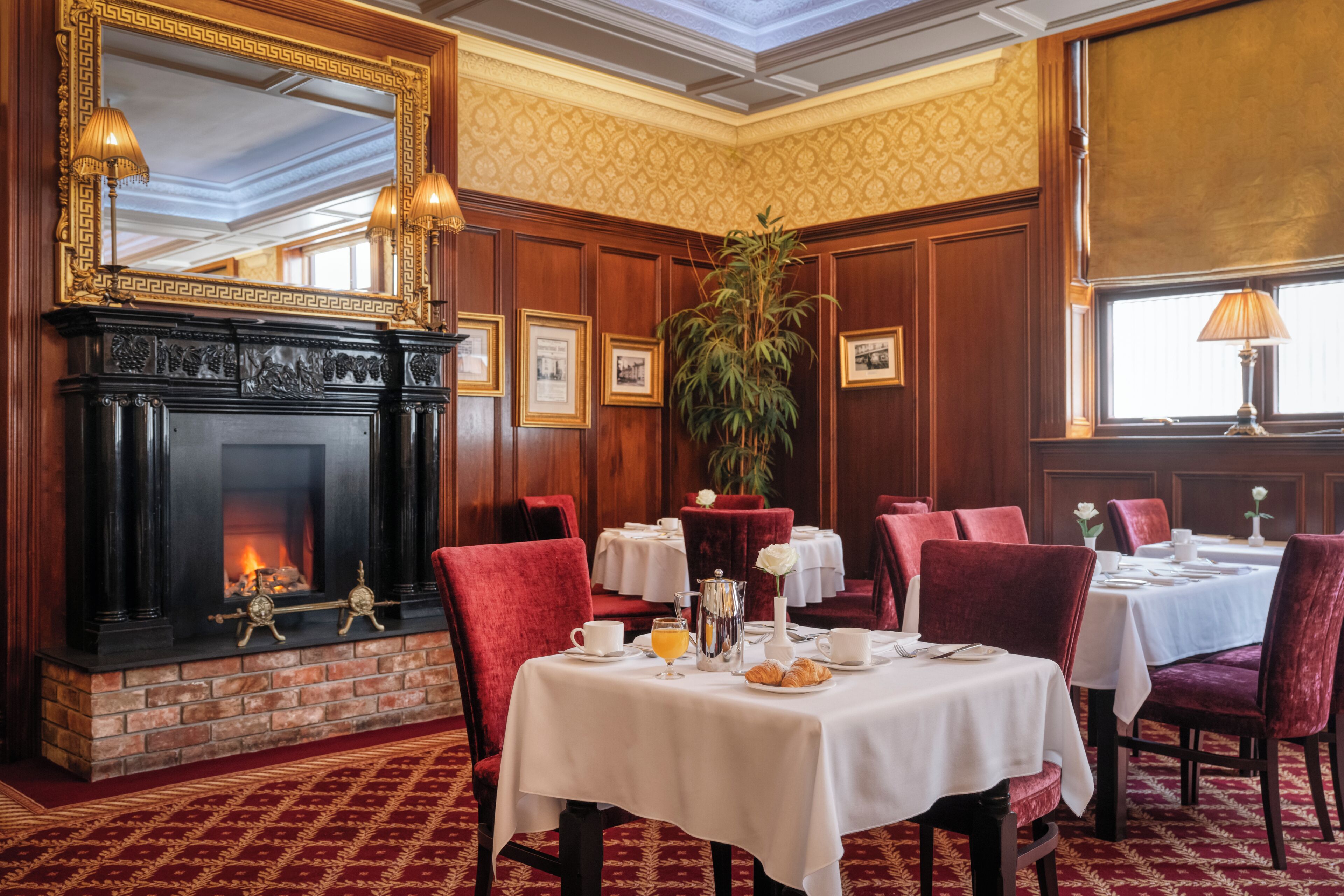 A restaurant with a fireplace, red upholstered chairs, white tablecloths, and framed pictures on the wall.