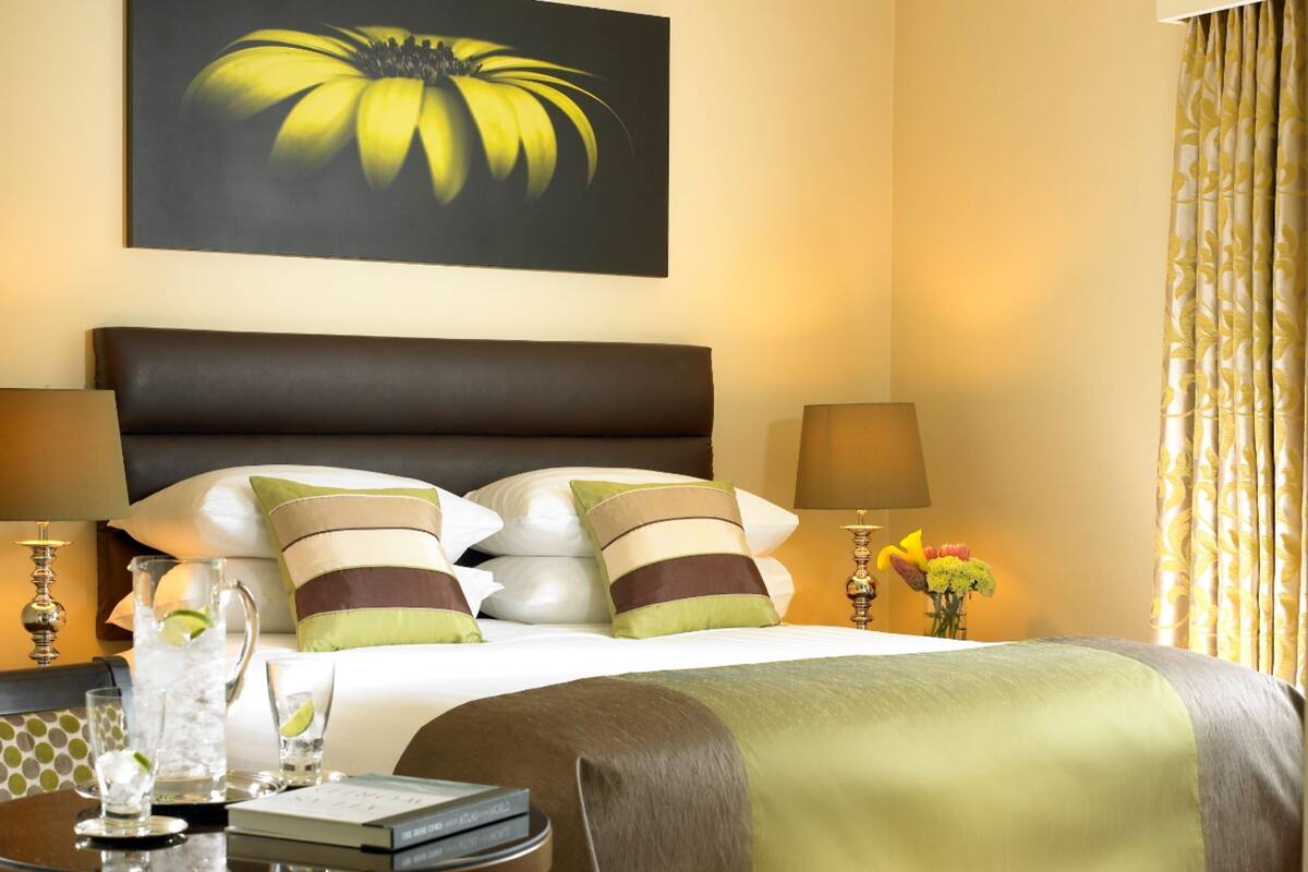 A hotel room with a large bed, two bedside lamps, a bedside table with a book and a glass of water, and a painting of a flower on the wall.