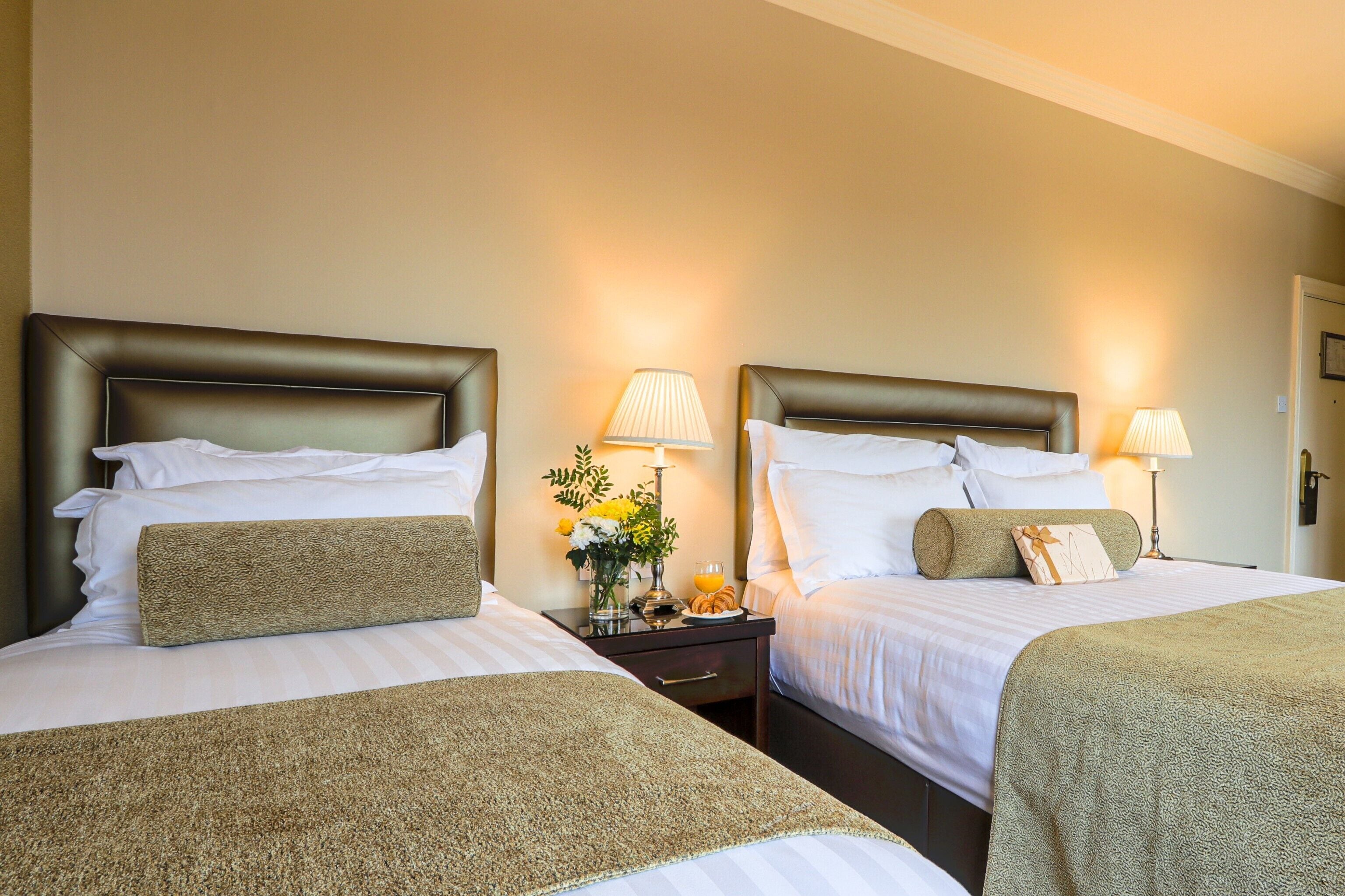 Habitación de hotel con dos camas, cada una con cabecera, mesitas de noche con lámparas y un jarrón con flores.