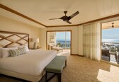 A hotel room with a large bed, a ceiling fan, a bench, a nightstand, a lamp, a view of the ocean through a balcony door, and a patterned carpet.